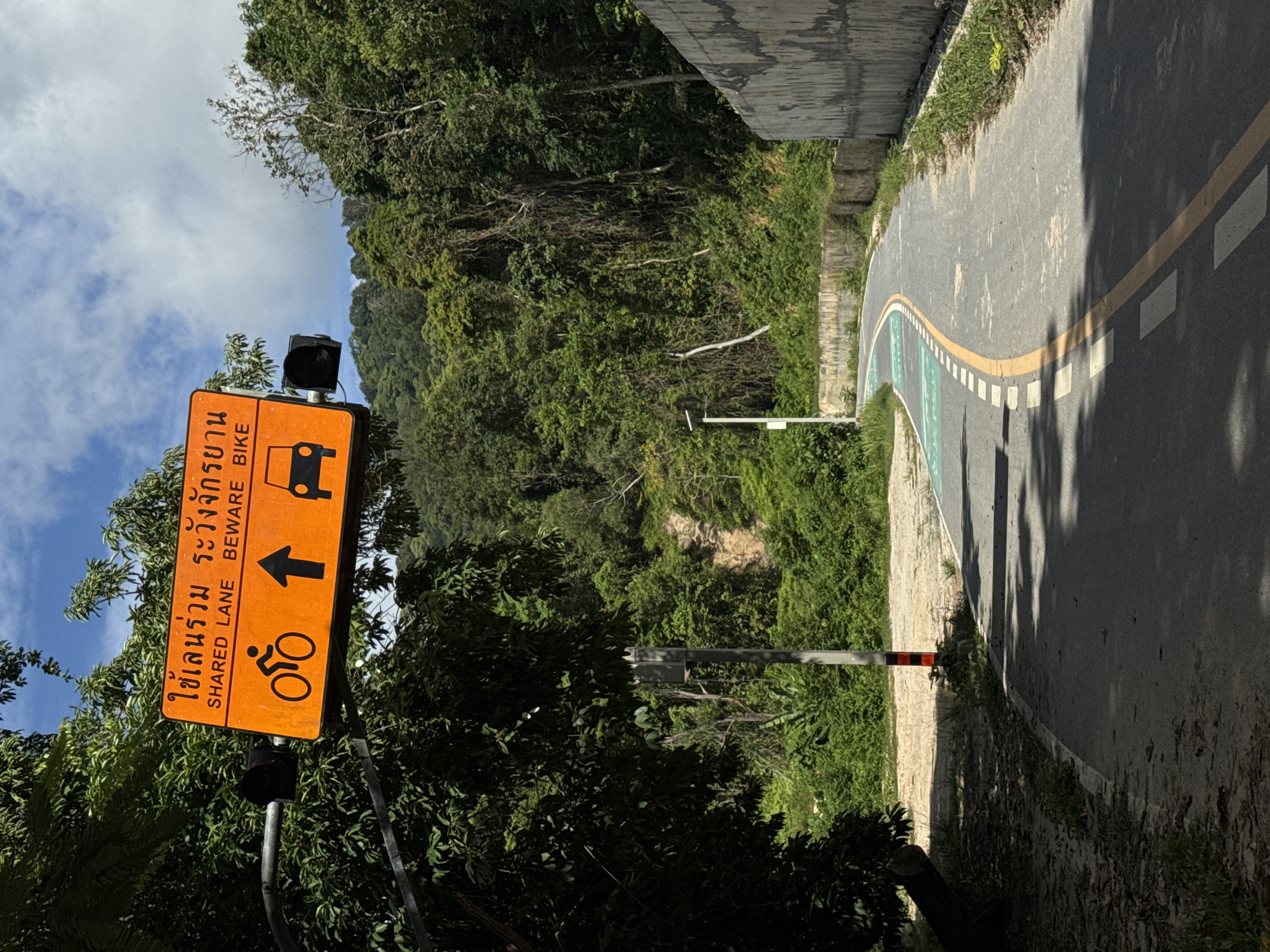 An orange shared lane sign with Thai text and English text "SHARED LANE BEWARE BIKE" is mounted on a metal pole over a paved path in Kathu, Thailand. The sign features icons of a bicycle, an upward-pointing arrow, and a car. The path curves to the right and is marked with white and yellow lines, and a green painted section. Lush green foliage and trees surround the path, with a concrete wall on the right side. A traffic light is also visible. The sky is partly cloudy.