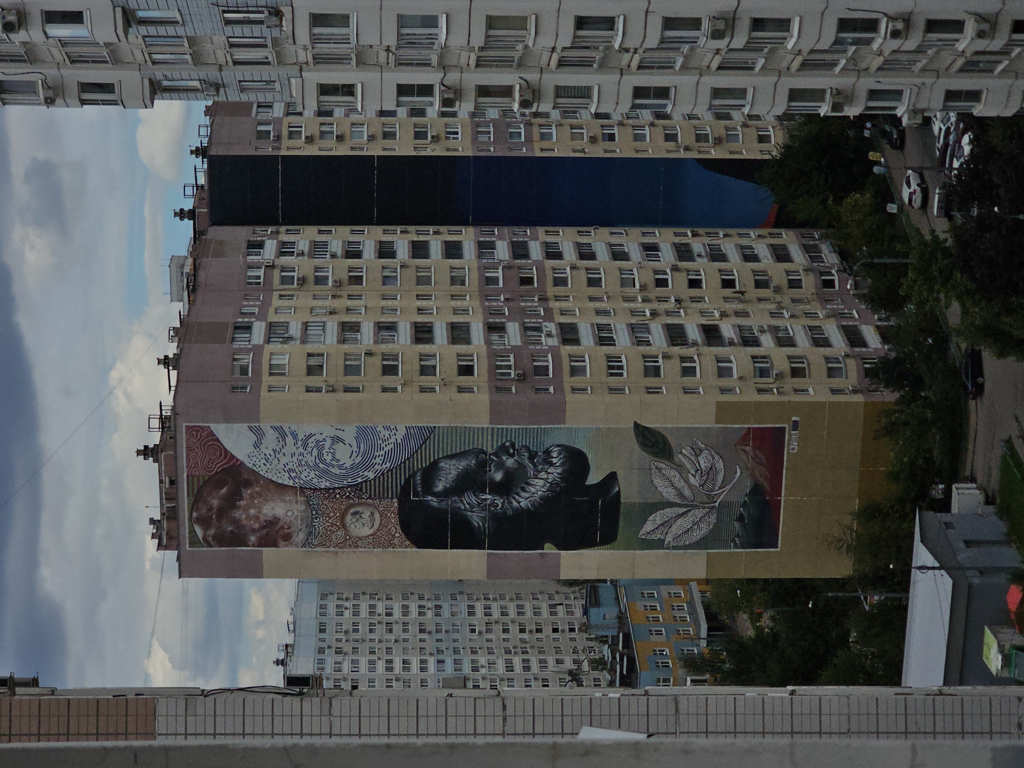 This is an aerial, medium-range shot of a residential area in Odintsovo, Russia, under a cloudy sky. Dominating the center is a tall, multi-story apartment building adorned with a large mural. The mural features a detailed portrait of a man's head in profile, rendered in black and white with a textured beard and flowing hair. Above the head, the mural depicts abstract circular patterns, including what appears to be a planet or moon, and swirling lines. Below the head, there are large, stylized leaves and a landscape with mountains. The building itself is painted in muted colors of yellow, pink, and beige, with rows of windows typical of Soviet-era apartment blocks.
To the left of the muraled building, another apartment building is visible, lighter in color and with a similar layout of windows. Further to the left, a segment of a gray, concrete building frame is in the foreground. To the right of the central building, a darker apartment building, seemingly painted black, breaks the colorful pattern of the mural. Adjacent to this, a lighter, gray-toned apartment building with curved balconies fills the right side of the frame.
At the base of the buildings, mature trees form a dense, green canopy. A street winds through the trees, with several parked cars visible, including white, black, and silver vehicles. A low-rise building, possibly a garage or utility structure, is situated near the bottom left of the image. The overall impression is one of urban development with artistic embellishments on a large scale.