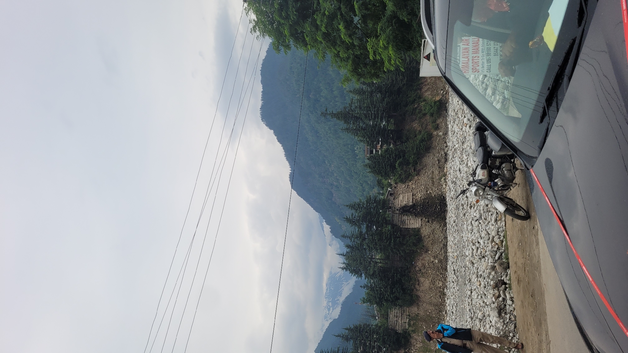 The image depicts a scenic view of the mountains and a riverbed in Vashisht, India. In the foreground, a black car is parked, partially obscuring the view. To the left, a person in a uniform resembling that of a police officer stands on a dirt path next to a rocky riverbed. A silver motorcycle is parked near the riverbank. The background features a dense forest of pine trees, a steep, rocky embankment, and imposing mountains, some of which are snow-capped. The sky is overcast with clouds. A sign on the car's windshield indicates "HIMALAYAN AIR SPORTS MANALI" with a contact number.
