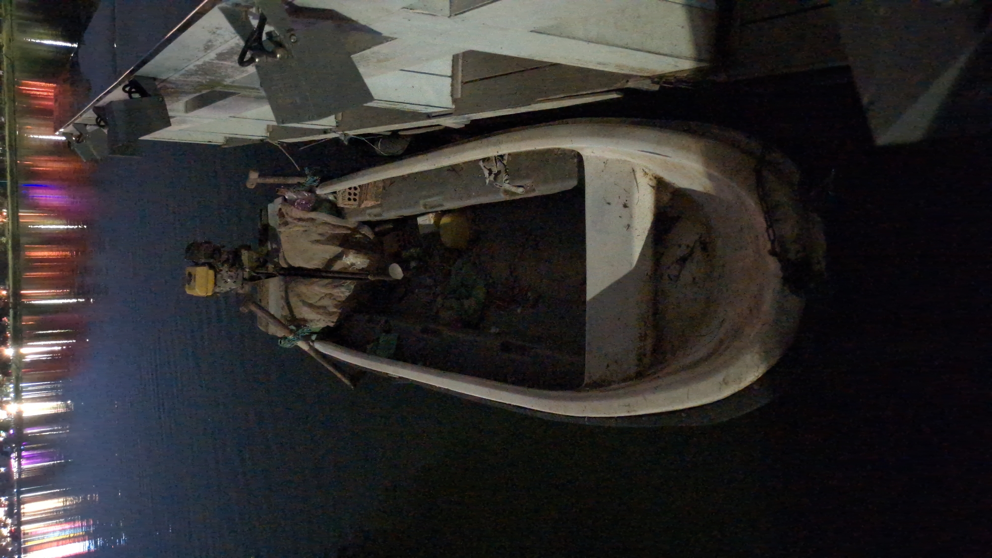 The image depicts a small boat on the water next to a larger structure. The boat is a light color and contains various objects inside, including a yellow object, bricks, and coverings. An outboard motor is mounted on the boat. The larger structure appears to be a dock or another boat. On the left side of the image, there are lights reflecting on the water, suggesting a city skyline at night. Considering the location requested, these might be lights of Da Lat reflected in Xuan Huong Lake. The surrounding water appears dark. The overall image quality is somewhat grainy, and the lighting is dim.