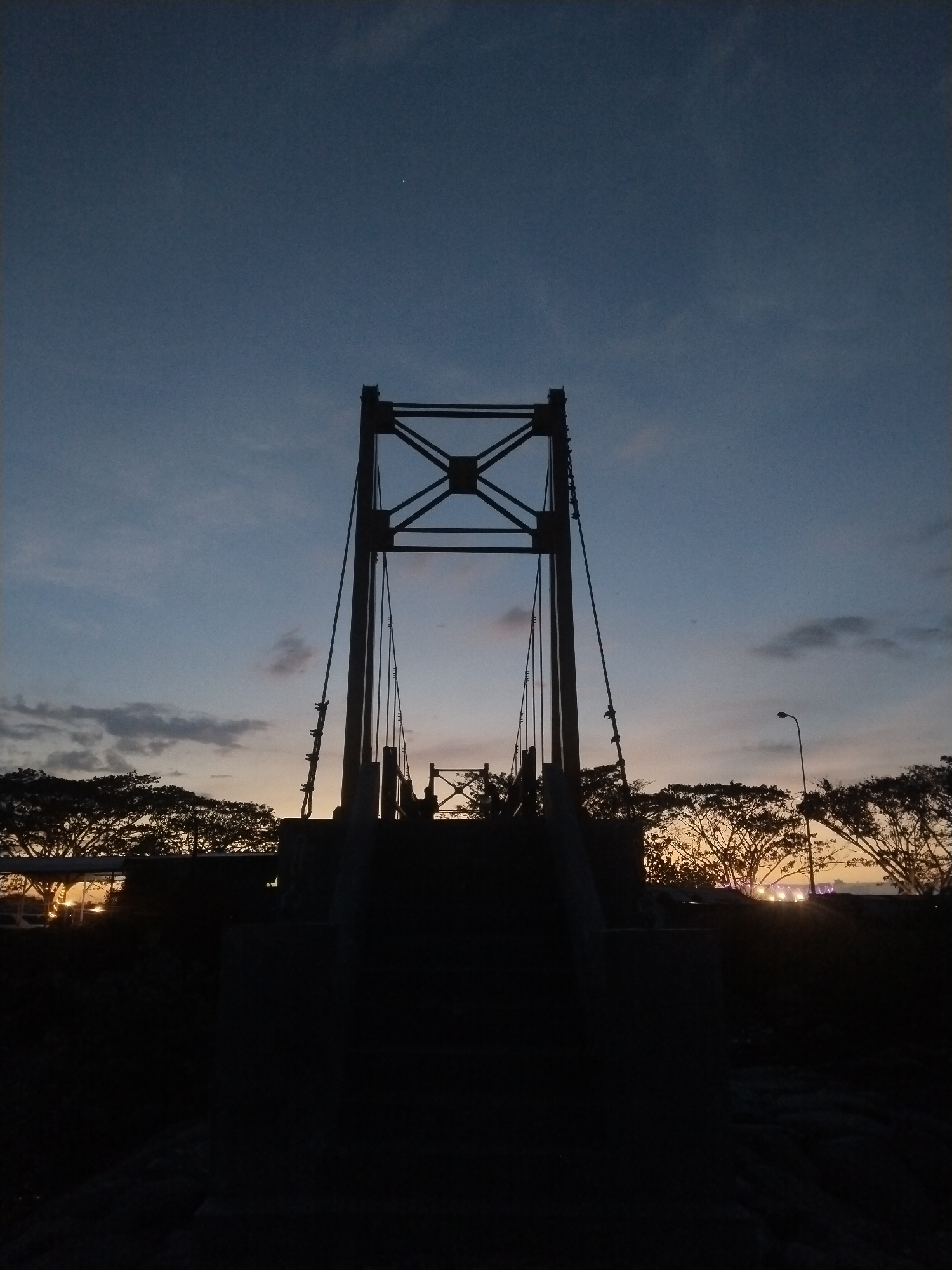 The image shows a bridge silhouette against a twilight sky in Lhokseumawe, Indonesia. The bridge's support structure is visible with cables extending downwards. Trees are silhouetted in the background, with some light visible through their branches. A street light is also visible among the trees on the right. The foreground is dark and obscures any details.