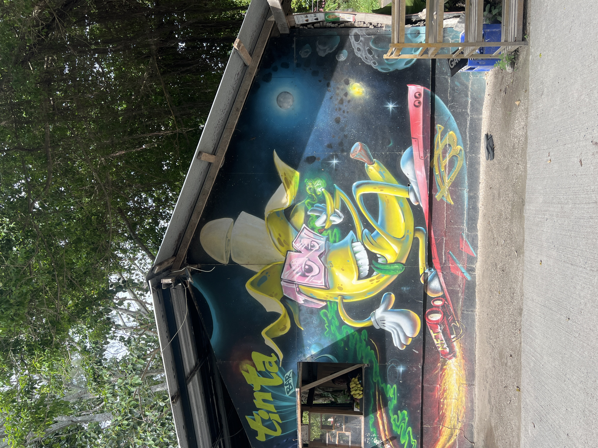 The image depicts a colorful mural painted on the side of a building in a tropical environment. The mural features a cartoonish yellow banana character wearing pink sunglasses and a cape, riding on a rocket that emits fiery exhaust. The background of the mural is a cosmic scene with stars, planets, and a moon. Above the mural, a corrugated metal roof is visible, supported by wooden beams. Lush green trees and foliage are seen in the background, casting shadows on the building. To the left of the mural, a small window is visible with a display of bananas inside. The foreground shows a concrete surface and a dirt area with a pair of flip-flops and a blue crate. The overall impression is a vibrant and whimsical artistic representation on the side of a casual establishment, likely a bar as indicated by the word "BAR" on the mural. This scene is located in Ko Pha Ngan, Thailand.