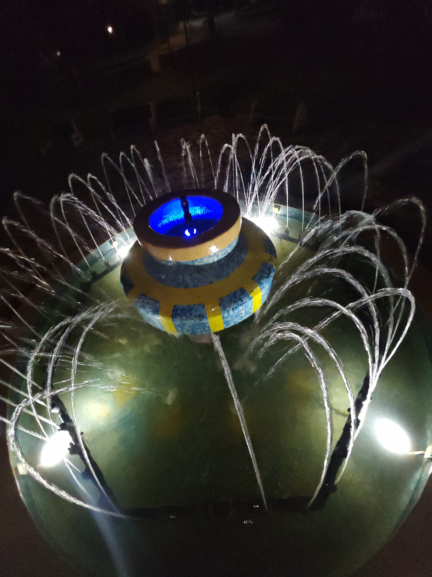 The image depicts an illuminated water fountain at night in Veliki Preslav, Bulgaria. The central feature is a tiered structure, cylindrical in form, adorned with blue and yellow mosaic tiles arranged in a band pattern. The top-most tier contains a concave basin emitting a bright blue light from within, with a small central water feature. Numerous thin water jets emanate in an arc pattern from the perimeter of the central tiled structure and also from the outer basin, creating a circular display. The main fountain basin is filled with dark green water, illuminated by several distinct white lights positioned around its outer edge, which highlight the ascending water streams. The surrounding environment is dark, with faint outlines of indistinct structures or benches visible in the distant background, suggesting an outdoor public space. No individuals or specific identifying landmarks beyond the fountain are discernable.