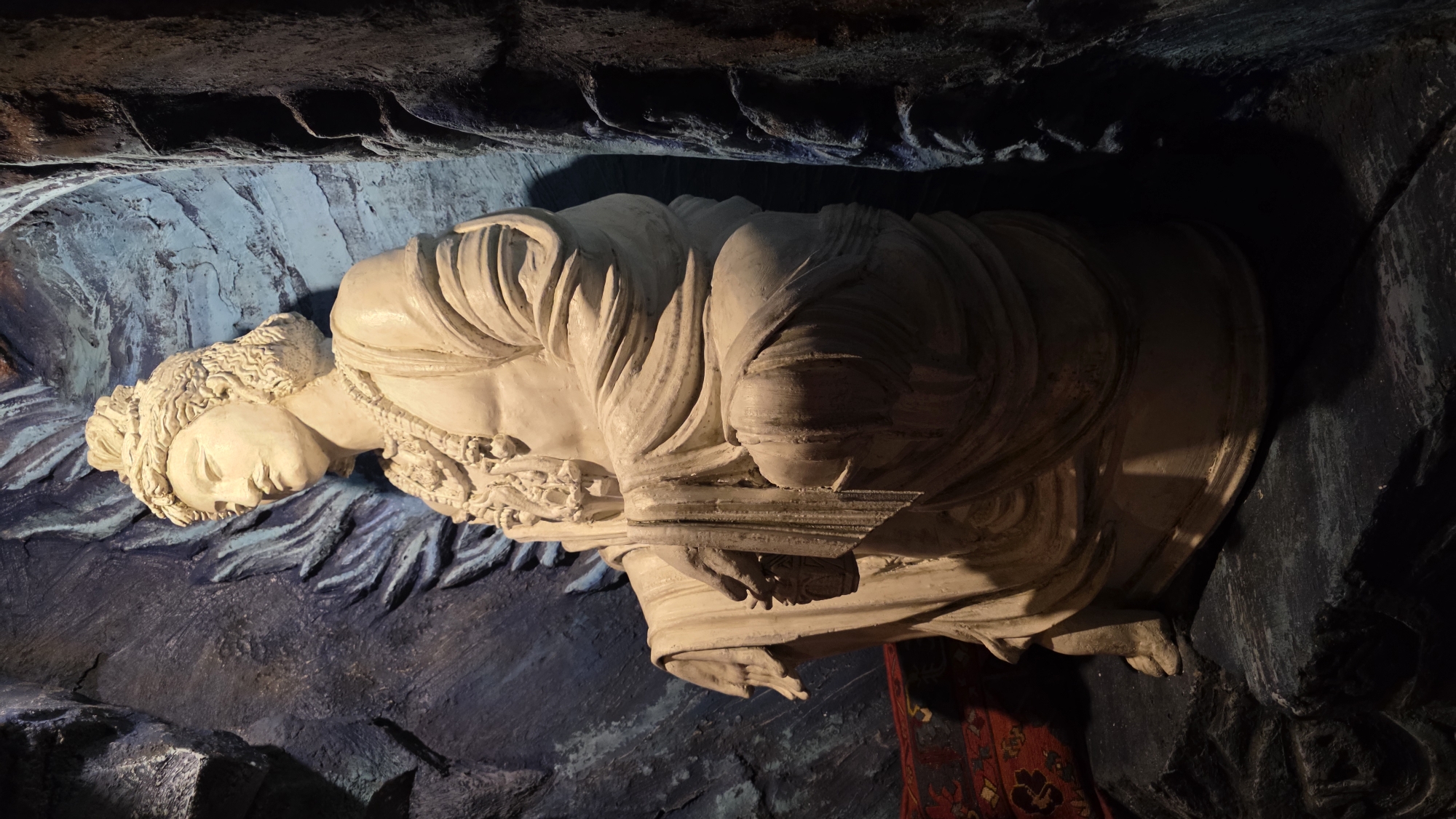 The image depicts a white statue of a woman, possibly a depiction of a reclining female figure from Greek mythology, housed in a cave-like environment. The statue's head is resting, and it appears to be draped in fabric. The surrounding environment is dark, resembling a cave or grotto with carved or sculpted elements. The statue is located in the grotto in front of the pavilion "Turkish Bath" in Catherine Park, Tsarskoye Selo, Saint Petersburg, Russia. A portion of a patterned rug is visible below the statue. The overall lighting emphasizes the statue's contours, giving it a three-dimensional appearance.