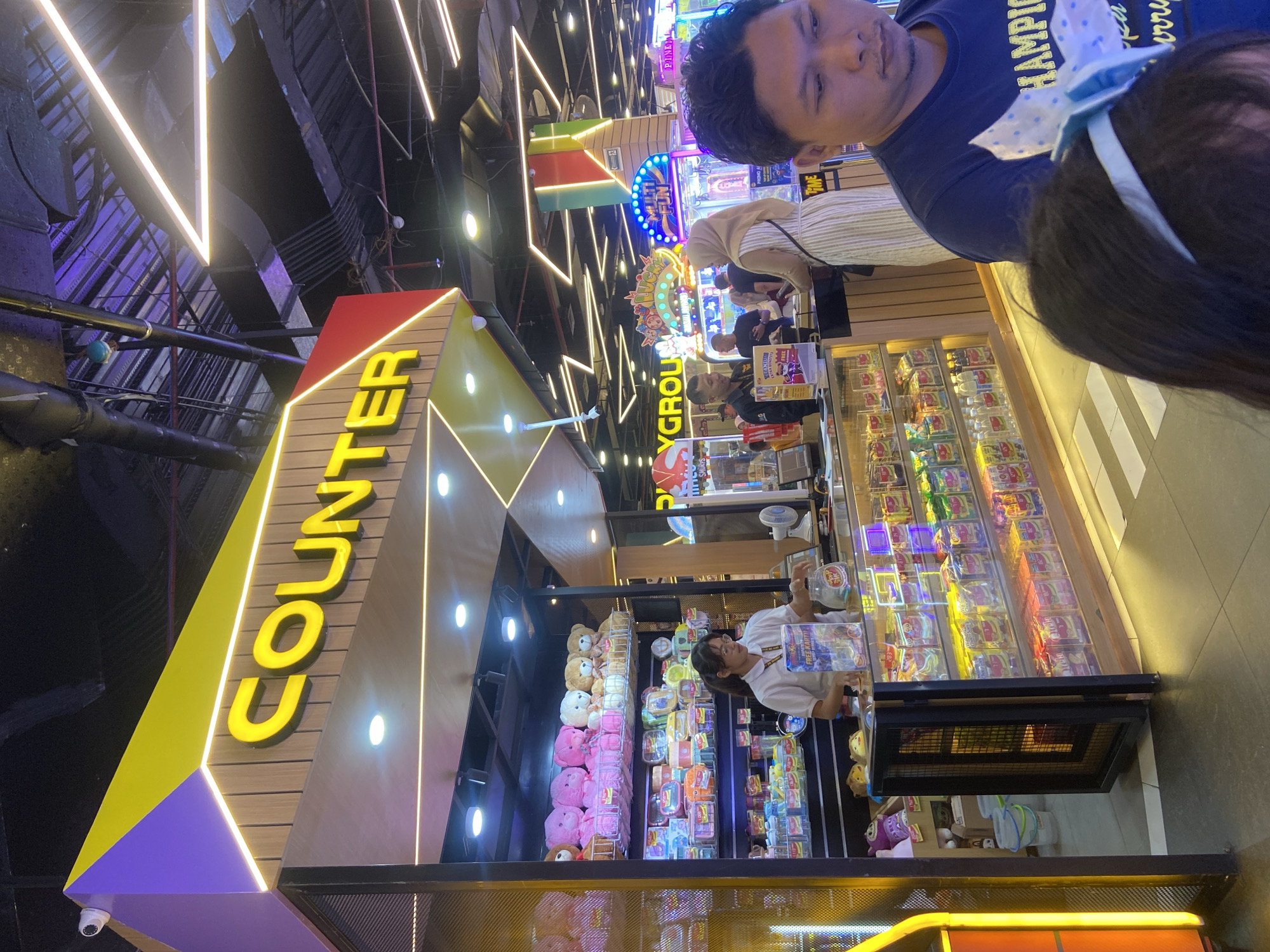 In Sidoarjo, Indonesia, this image captures a brightly lit arcade with a prominent "COUNTER" sign. A female employee wearing a white shirt stands behind a counter filled with plush toys and various packaged goods. Adjacent to her, a male customer in a dark blue shirt is visible in the foreground. In the background, other arcade machines and patrons can be seen, illuminated by colorful neon lights. The overall atmosphere suggests a lively entertainment venue.