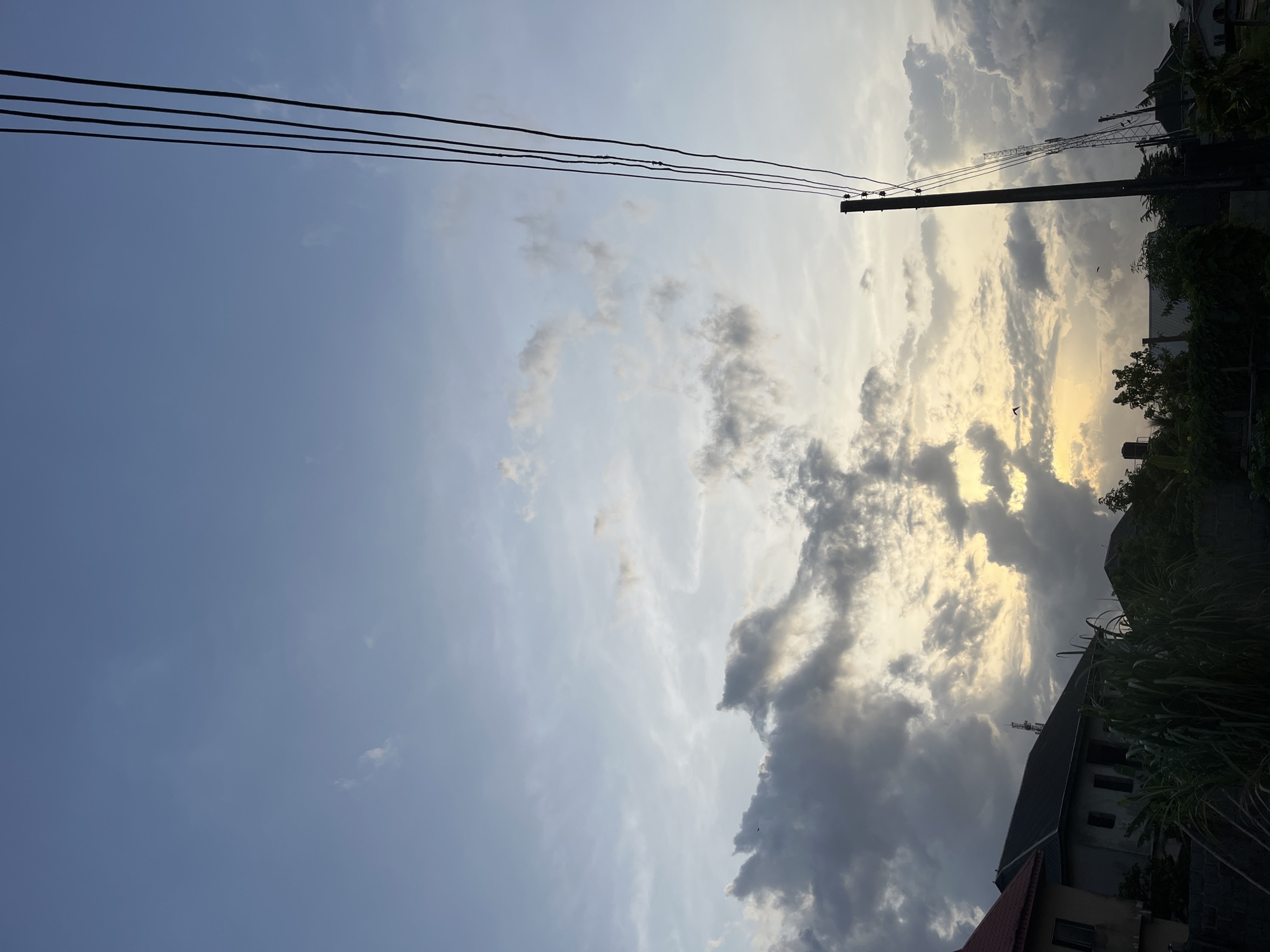 The image shows a sky with scattered clouds, ranging from light wisps to dark, dense formations, suggesting either a sunrise or sunset. Sunlight is breaking through the clouds in the upper right portion of the frame. Power lines run across the upper part of the image, and a utility pole is visible on the right side. Vegetation and parts of buildings are visible on the right and bottom, indicating a residential or suburban setting. There is no information available about the location of the city.