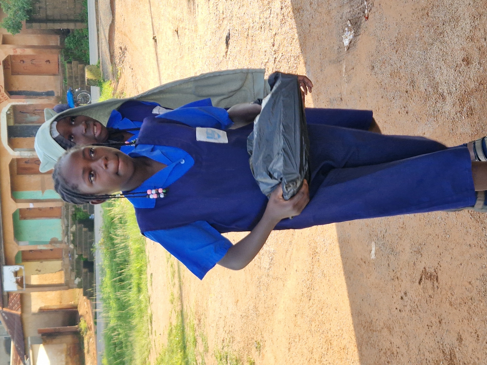 Two young girls, appearing to be of African ethnicity, stand in a dirt-floored outdoor area. The girl in the foreground is wearing a royal blue uniform, possibly a school uniform, consisting of a short-sleeved shirt and a jumper or dress. She has dark hair braided with pink and white beads and is holding a black plastic bag. To her right, a second girl is also visible, wearing a similar royal blue uniform and a light green head covering that drapes over her shoulders. The background shows buildings with corrugated metal roofs and stucco walls, some with doorways and windows. Vegetation, including grass, is visible to the left of the girls. The ground is covered in reddish-brown dirt and small stones. A portion of a motorcycle is visible in the distance, near a building. The overall scene suggests an environment in a rural or semi-urban setting, consistent with a location in Kabong, Nigeria.