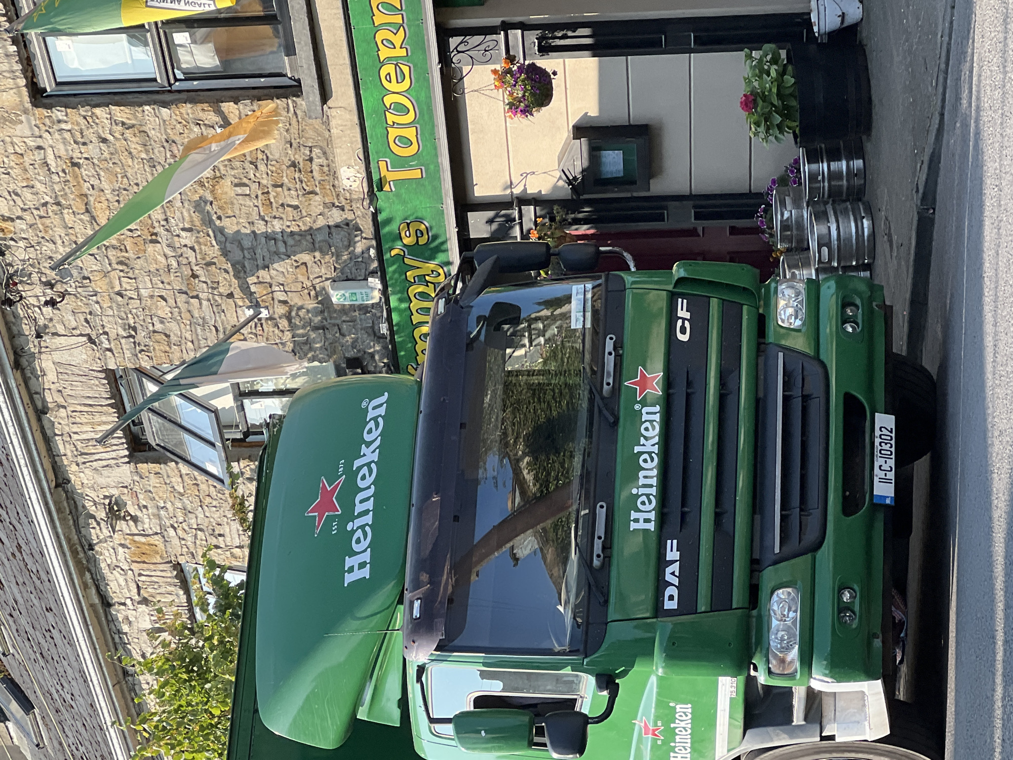 A green Heineken truck is parked on a street in front of a stone building. The building has a sign that reads "Tommy's Tavern". There are Irish flags hanging outside the tavern. The truck has "Heineken" and "DAF" logos on it. Several metal beer kegs are stacked on the sidewalk next to the truck. There are also potted plants outside the tavern. The scene is well-lit, suggesting it is daytime. The license plate on the truck is "II-C-10302". The location is Mountcharles, Ireland.
