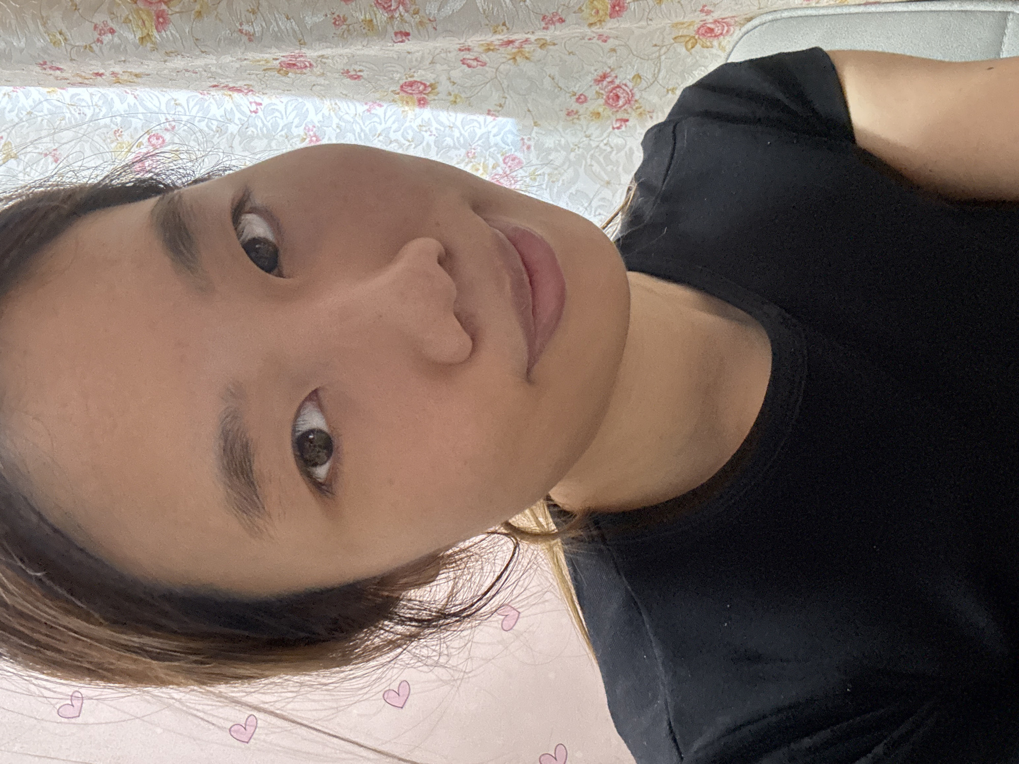 A close-up selfie of an East Asian woman is taken from a slightly low angle. She has light brown hair pulled back, and her face is partially illuminated by natural light coming from her right. She is wearing a plain black t-shirt. Behind her, a light-colored curtain with a floral pattern is visible on the right side of the frame. On the left, a pink background with three small, light pink heart illustrations is present. The scene appears to be indoors. The location is not specified in the image or its metadata.
