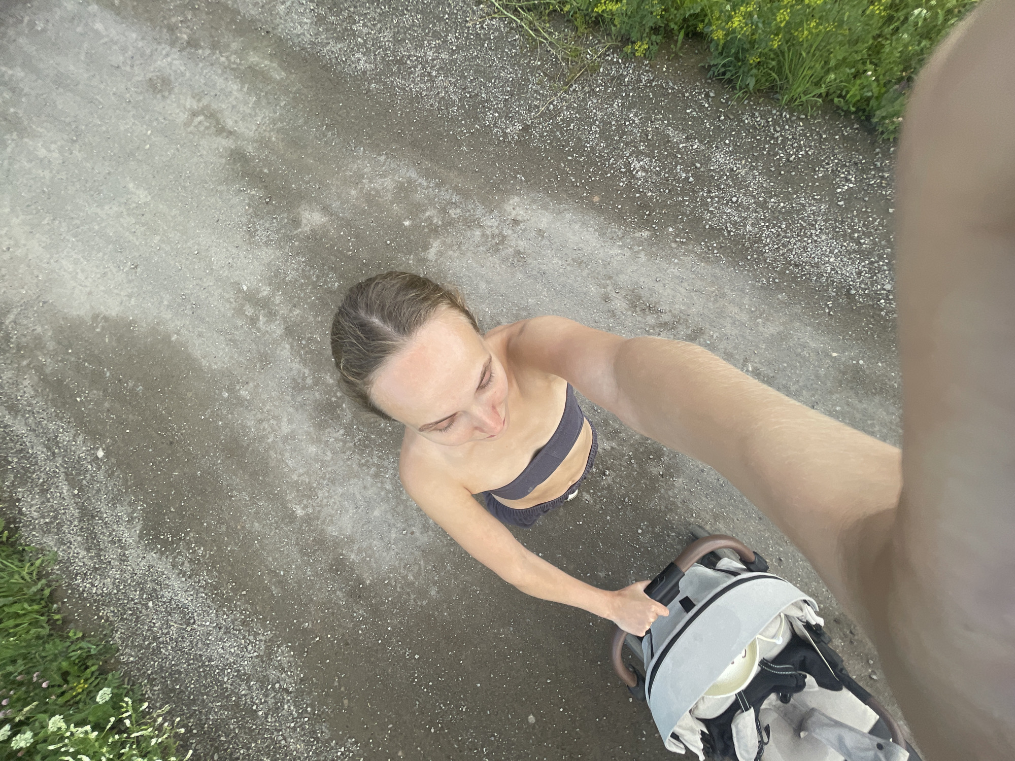 An overhead shot captures a woman from the shoulders up, pushing a baby stroller along a gravel path. She is looking down and to her right, with her hair pulled back. She wears a dark-colored bandeau top and dark shorts. The stroller is light gray and white. To the left of the path, there is green grass with some small pink and white flowers. To the right of the path, there is more green vegetation with yellow flowers. The ground is predominantly composed of gray gravel with some larger stones. The scene appears to be outdoors, possibly in a park or a less-developed area, given the natural vegetation and gravel path. The photograph is taken from a high angle, looking down at the woman and the stroller. The location is identified as Podshivalovo, Russia.