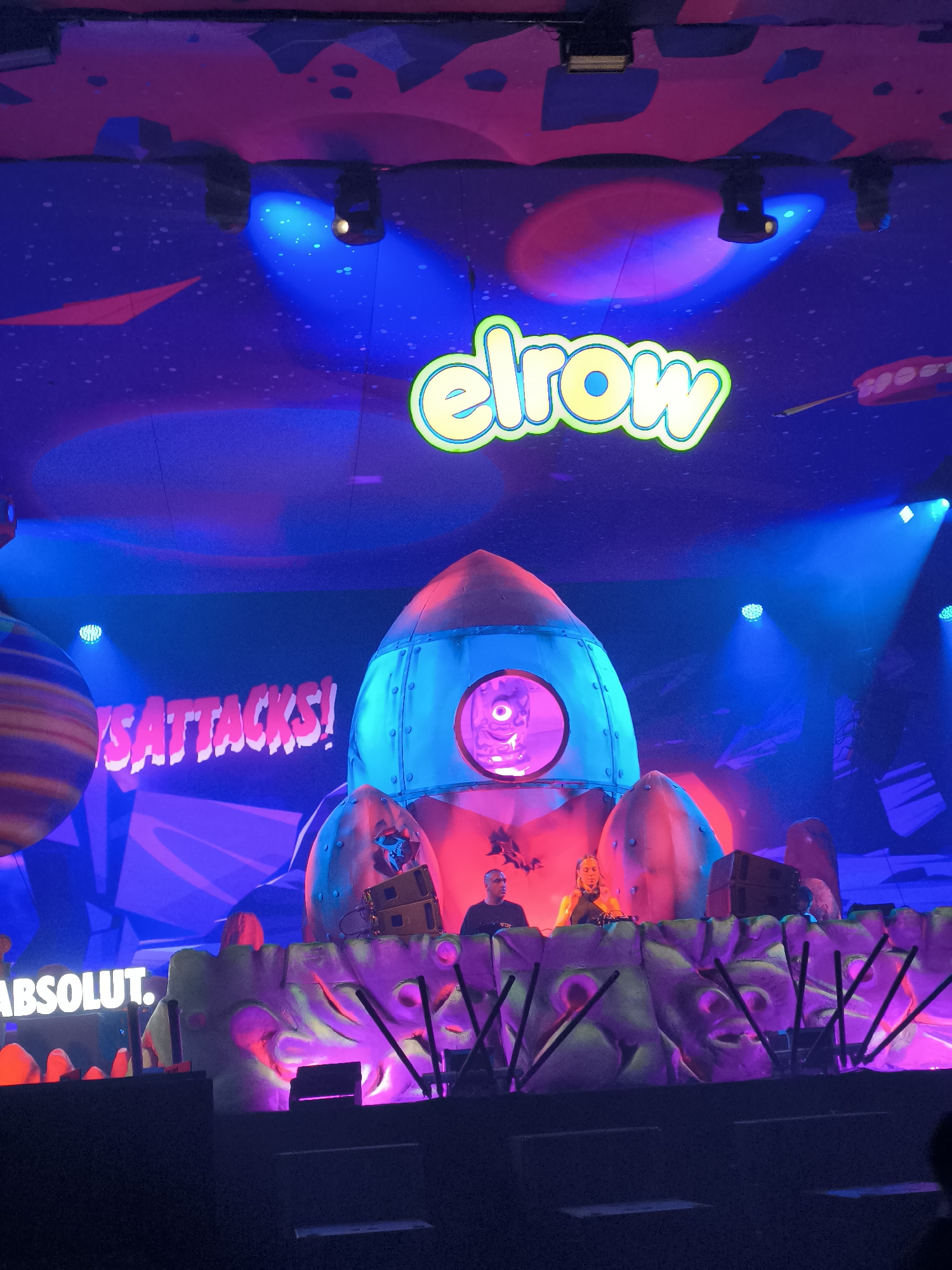 A stage, likely in Sofia, Bulgaria based on the background details, is decorated with a large cartoonish rocket and a large "elrow" sign. The words "SATtACKS!" are also visible on the set. The backdrop is a stylized space scene. On stage, two individuals, possibly DJs, are positioned behind a decorative wall with embedded facial features. An Absolut Vodka sign is visible on the side of the stage. Stage lighting is prominent.