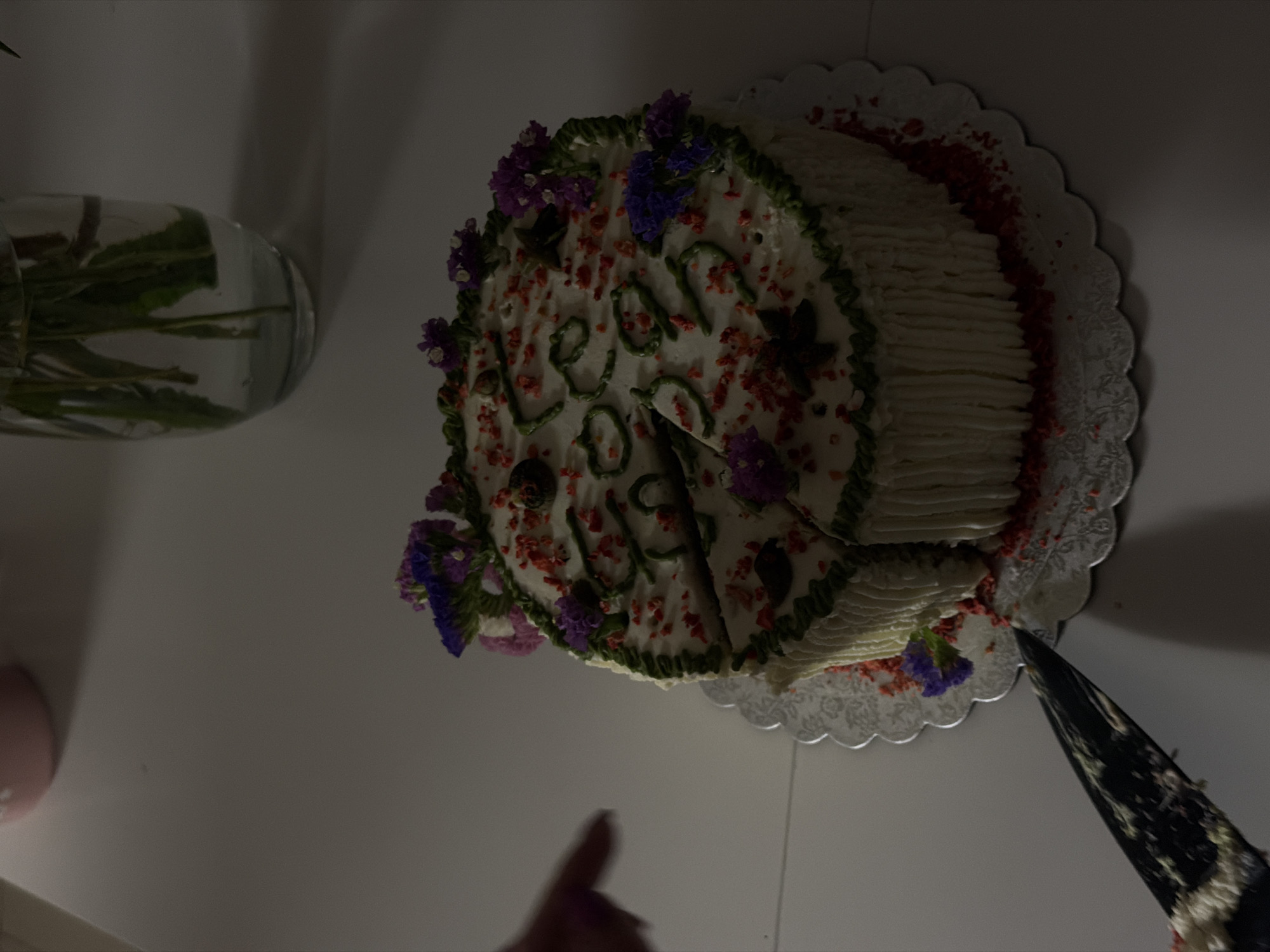 A cake is on a table, with green frosting on top forming the numbers "2022". The cake is decorated with red sprinkles and small purple and pink flowers. The cake has been cut into several slices, and a knife is positioned to cut another slice. A person's hand is visible reaching towards the cake. In the background, a glass vase with flowers and a pink object are visible. The scene appears to be indoors, on a table with a white surface. The location is Vračar Urban Municipality, Serbia.
