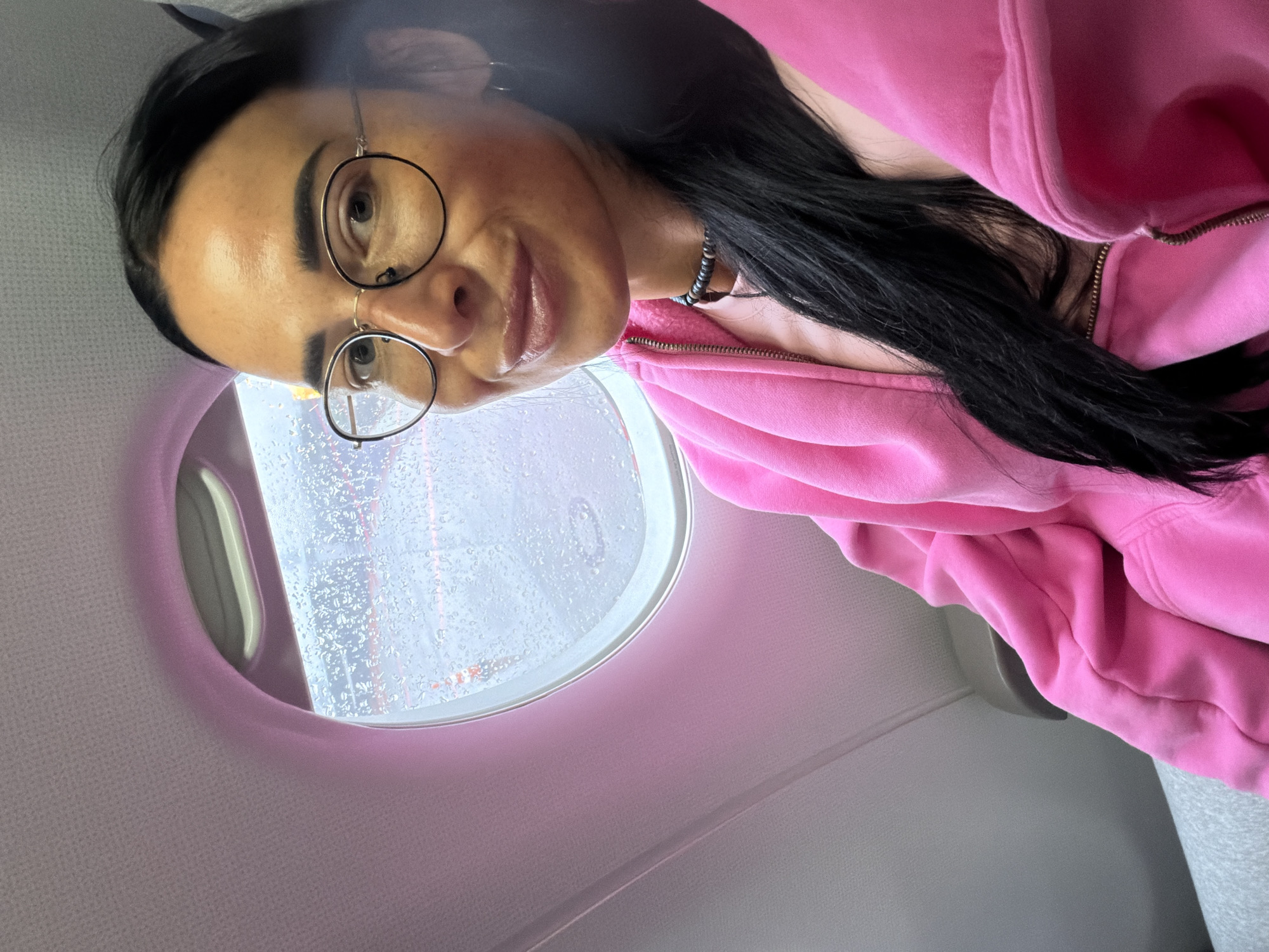 A woman wearing round glasses and a pink zip-up hoodie is seated next to an airplane window, which is covered in raindrops. Her dark hair falls over the hoodie. A necklace is visible at her throat. The setting appears to be inside an airplane, with the window providing a view of the exterior in rainy conditions. The location in the city of Khimki, Russia cannot be determined from the image.