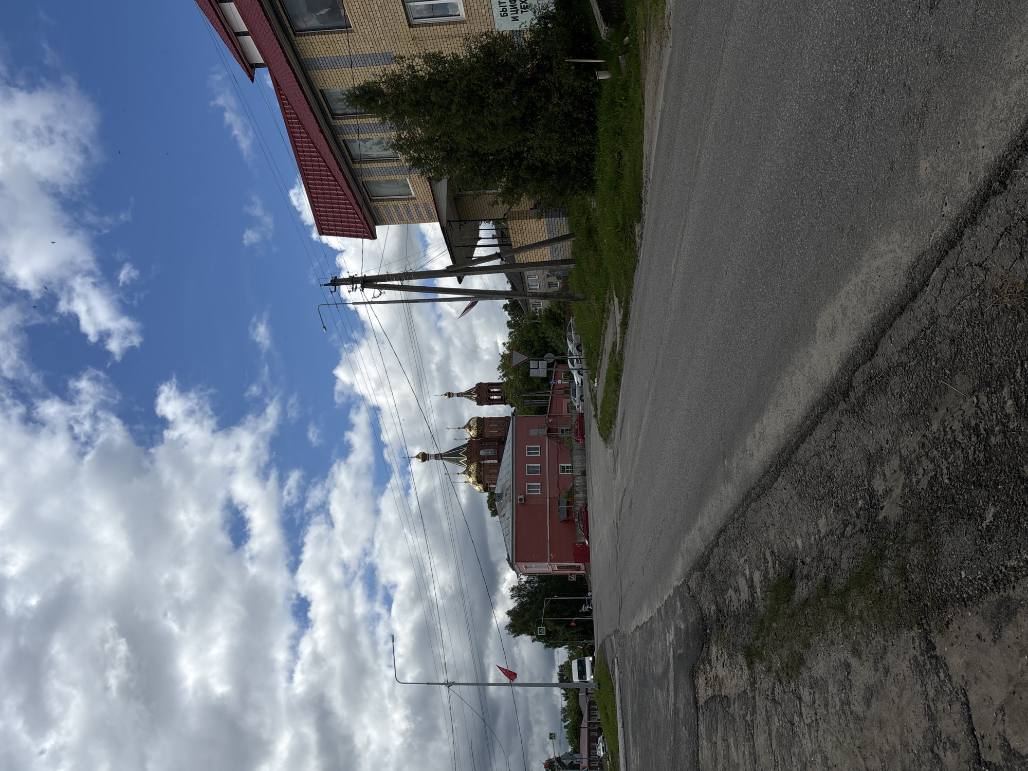 The image captures a street scene in Красные Баки, Russia, under a bright, partly cloudy sky. The perspective is looking down a road with buildings lining both sides.
On the left side of the frame, a multi-story building with a reddish-brown tiled roof and light-colored brick walls is visible. Its facade features partial Cyrillic text: "БЫТОВАЯ И ЦИФРОВАЯ ТЕХ..." which likely translates to "Household and Digital Appliances/Technology." Further along this side, an Orthodox church with reddish-orange walls and several golden onion domes stands out, indicative of Russian architecture. Adjacent to the church is another red-painted building.
An asphalt road runs from the foreground into the distance on the right, bordered by a curb and some grassy patches. Utility poles with numerous overhead wires and a street lamp are prominent along the road, and a small red flag is attached to one of these poles. Traffic signs are visible further down the road, and several vehicles, including a white van or truck, are present in the background. The sky is bright blue with scattered white cumulus clouds, and a few small birds are visible flying.
The scene depicts a calm, daytime atmosphere with no specific event occurring, simply a slice of daily life in a Russian town.