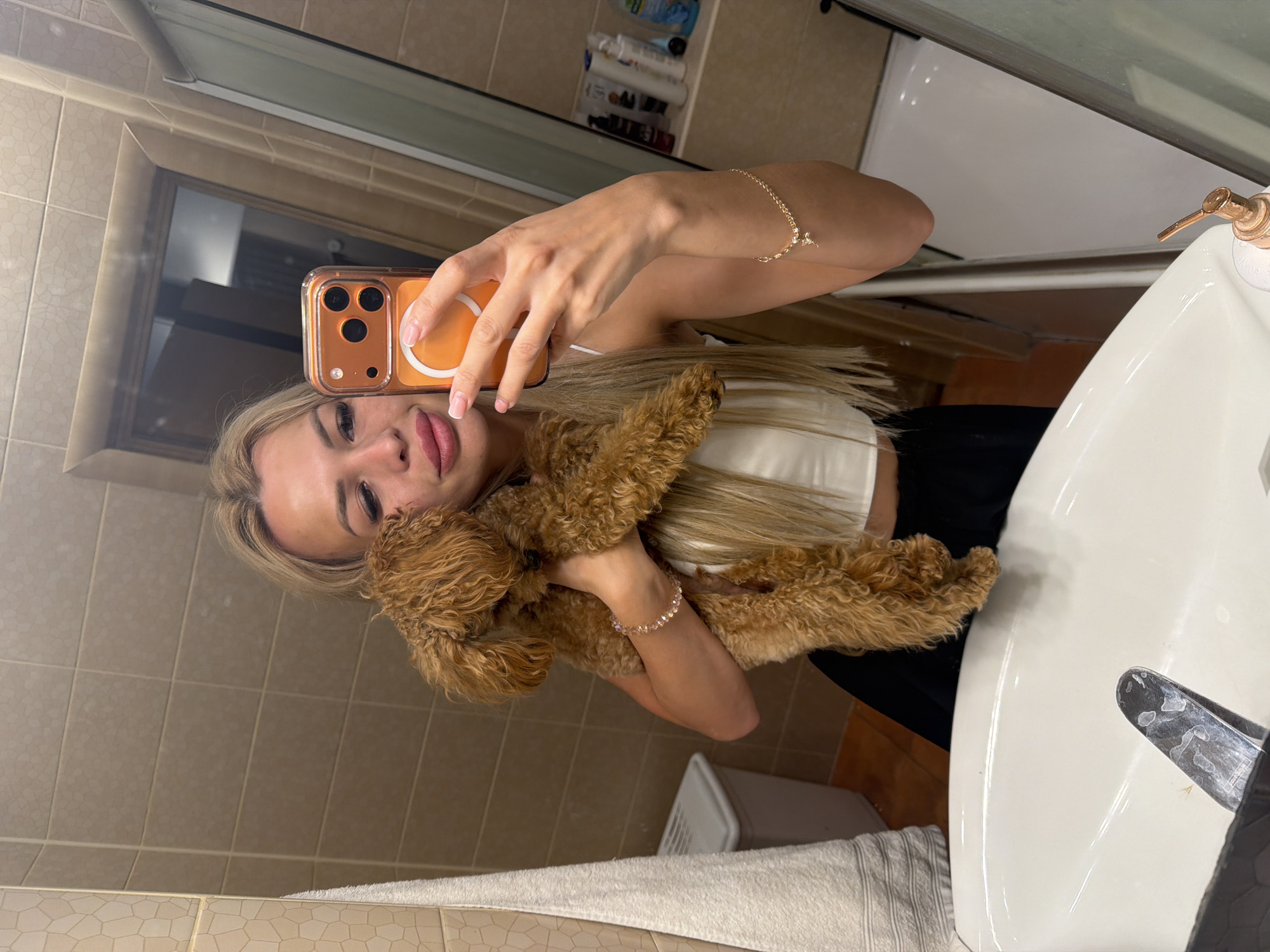 A young woman with blonde hair is taking a mirror selfie in a bathroom. She is holding a small, fluffy brown dog close to her chest. The woman is wearing a white crop top and a black skirt. She has on a delicate bracelet on her left wrist. The dog is a poodle or poodle mix.
The bathroom has tiled walls and a white sink. A soap dispenser is visible on the edge of the sink. In the background, there are shelves with various bottles and toiletries. The lighting suggests it is daytime, possibly morning or afternoon, as there is natural light coming from somewhere, illuminating the scene. The overall mood is casual and affectionate, as indicated by the way the woman is holding the dog.
There is no visible text in the image.
The location context is Kyiv, Ukraine.