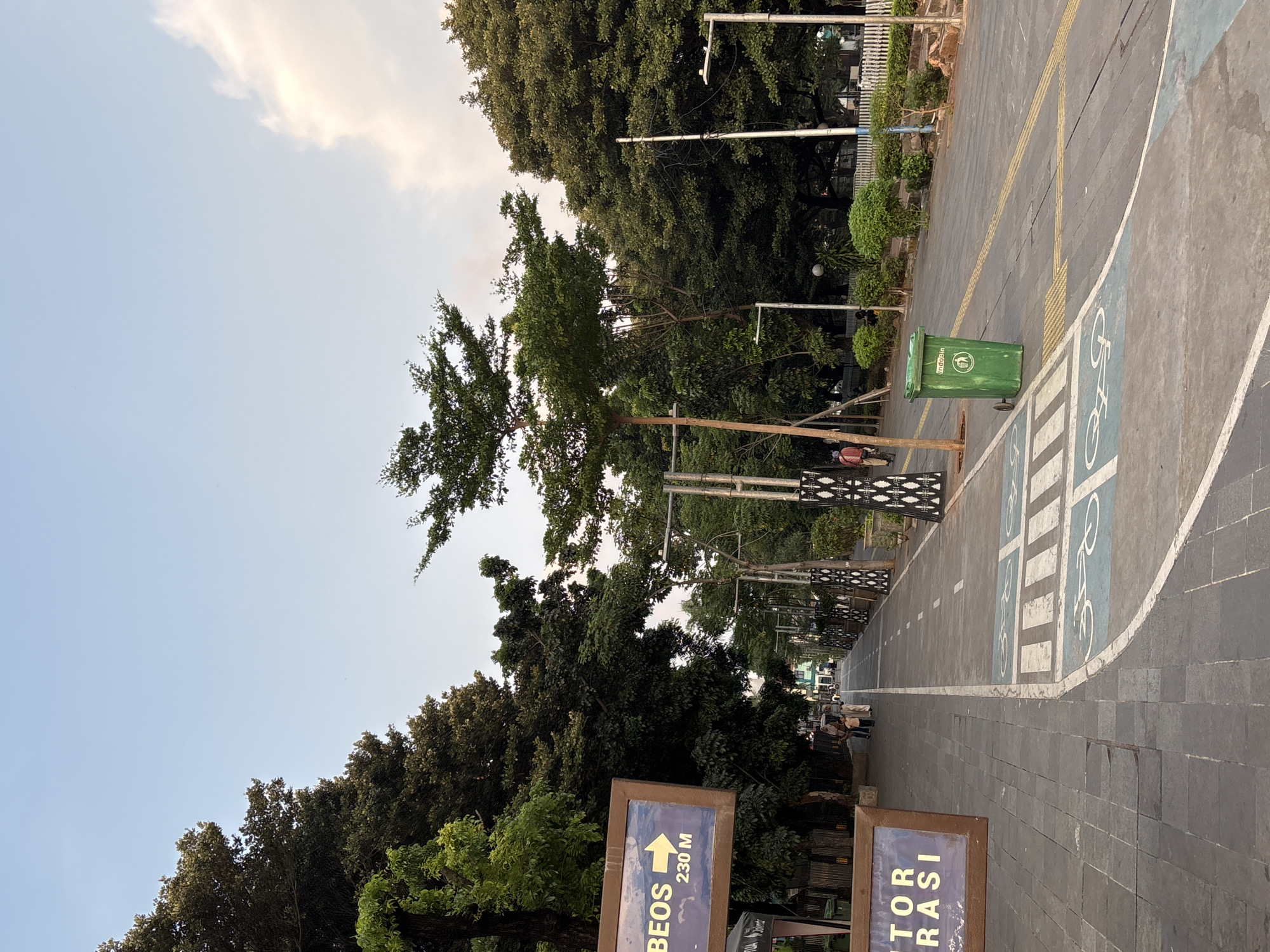 The image depicts a paved pathway lined with trees, likely in a park or urban green space. On the left, a sign with blue lettering and an arrow pointing right reads "BEOS 230 M." Below it, another sign partially visible says "TOR ERASI." A green trash bin is situated on the right side of the pathway. The pathway is marked with white lines and symbols indicating a bike lane. Several trees, some tall and slender and others with dense foliage, create a canopy overhead. In the background, people can be seen walking along the path. The sky above is a pale blue with a few wispy clouds. The overall scene suggests a well-maintained outdoor area in a city. This location is in the city of West Jakarta, Indonesia.