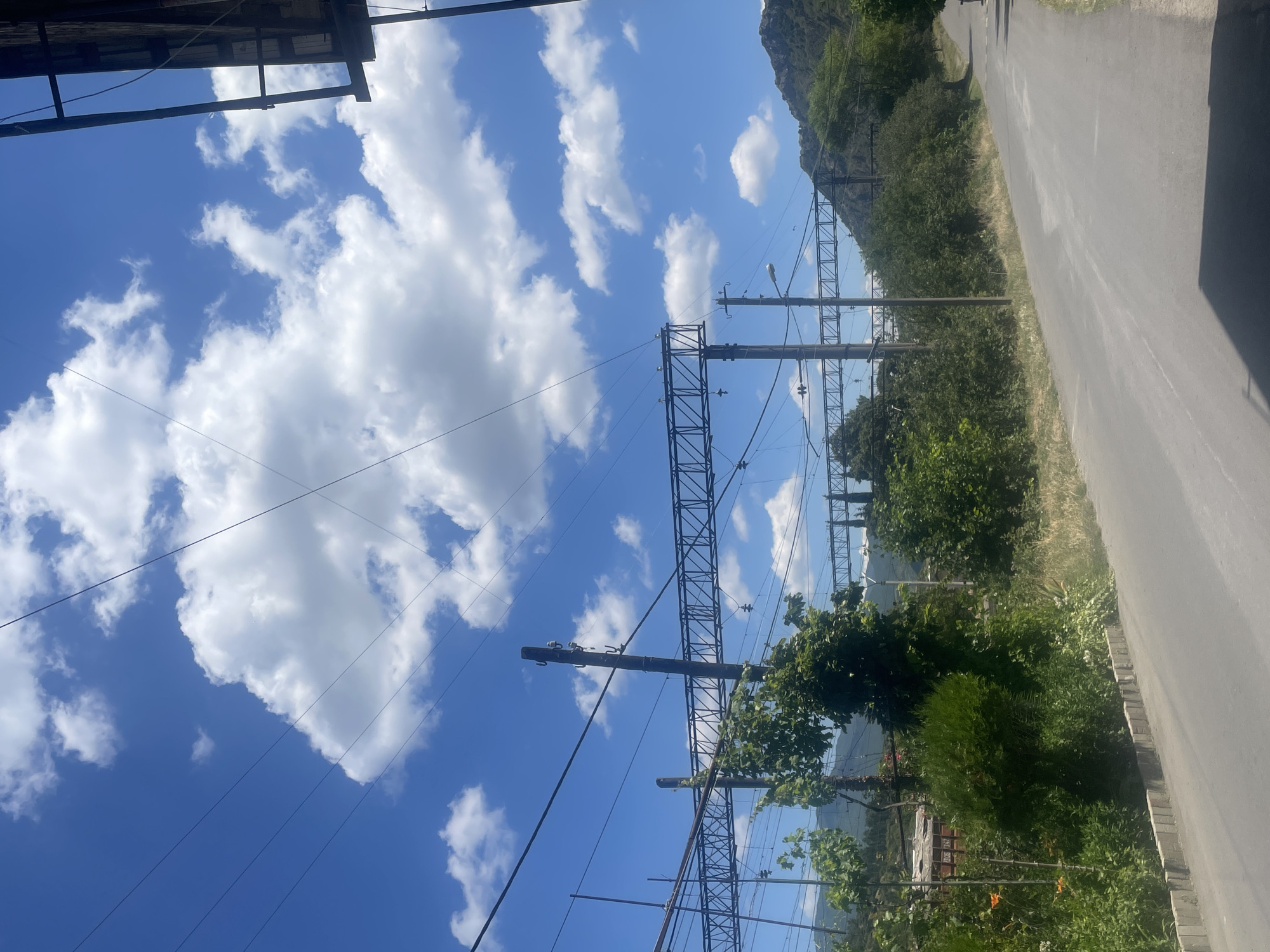 The image depicts a bright, sunny day in Mtskheta, Georgia, with a clear blue sky dotted with fluffy white clouds. The scene is dominated by a power line pylon and overhead wires, stretching across the frame. These lines traverse through lush green trees and vegetation that line a paved road, which occupies the right side of the image. In the background, a large, rocky hill is visible, partially covered by greenery. The perspective is upward-looking, emphasizing the vastness of the sky and the scale of the infrastructure against the natural landscape. The overall impression is one of a tranquil, rural or semi-rural environment, possibly near a railway line, given the presence of the power lines. There is no visible text in the image.