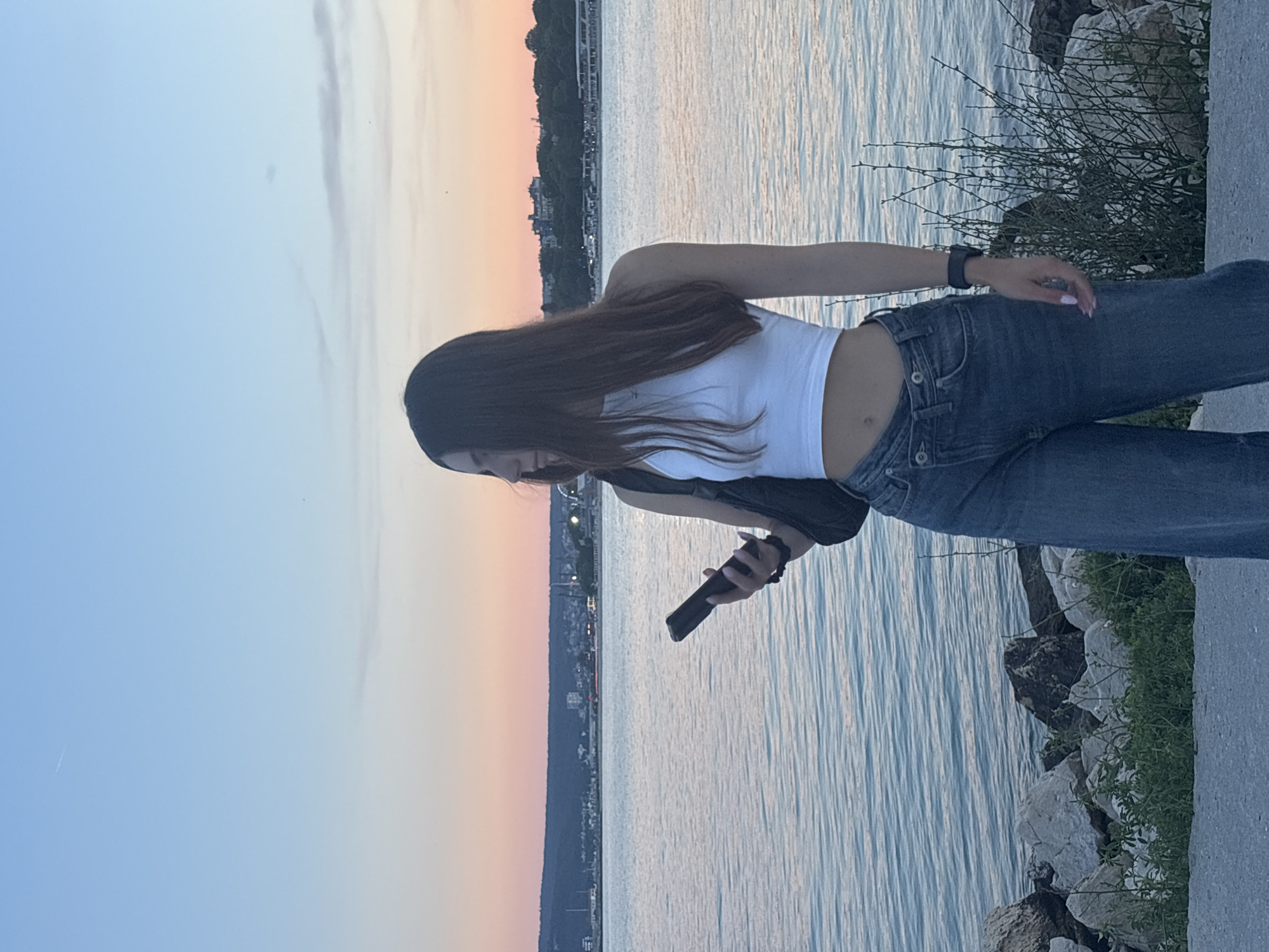 A young woman with long dark hair stands on a paved area by the sea in Varna, Bulgaria, holding a smartphone in her right hand. She is wearing a white crop top and blue denim jeans. The sea is calm and reflects the colors of the sunset sky, which transitions from pale blue at the top to pink and orange near the horizon. Across the water, a distant cityscape with buildings and lights is visible, nestled against a dark, tree-covered hill. The foreground features a rocky shoreline with scattered green vegetation. The overall scene depicts a serene evening by the sea in Varna.