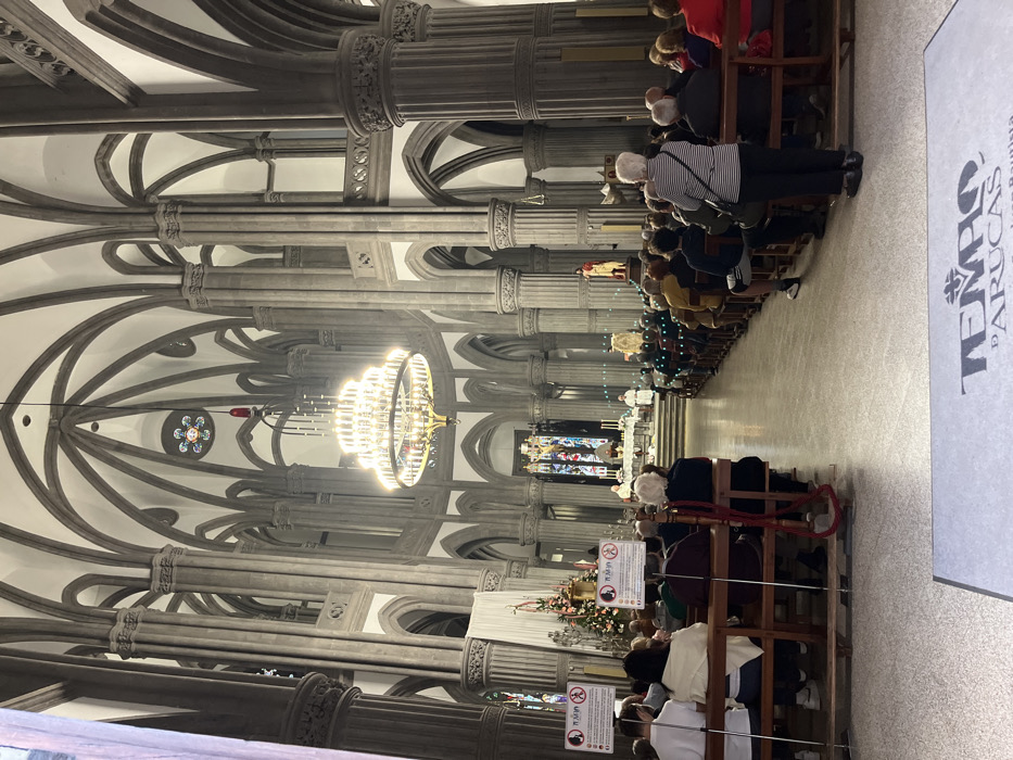 The image shows the interior of the Templo de Arucas, a church located in the city of Arucas, Gran Canaria, Spain (latitude: 28.119, longitude: -15.523). The church's interior is dominated by tall grey columns, intricate arches, and a large chandelier hanging from the high vaulted ceiling. The floor is tiled and there are several rows of pews filled with people attending a service. At the far end of the church, a small group of people are standing in front of an ornate altar decorated with candles. There are several people standing at the front, including two people in white robes. The church is decorated with Christmas trees and lights, and the walls are decorated with various religious artwork.
