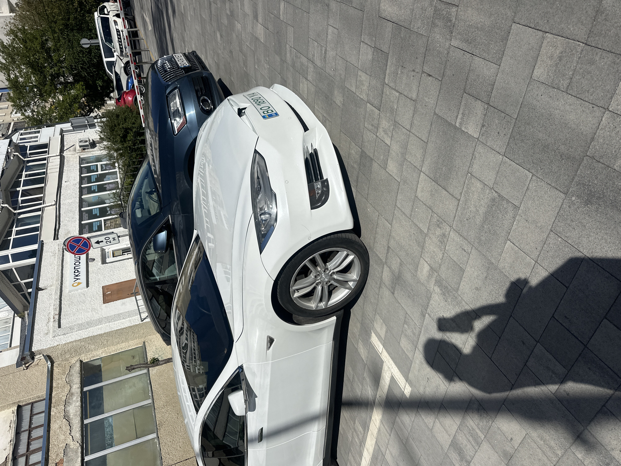 This is a medium shot, eye-level, exterior, sunny day photograph taken at the Ukrposhta (Ukrainian Postal Service) branch located at 13B Heroes of Maidan Street in Ternopil, Ukraine. In the foreground, a white Tesla Model S is parked, facing right. Behind it, a dark blue Audi Q5 is also parked. The ground is paved with grey square tiles. A shadow of a person holding a smartphone, presumably taking the photo, is cast on the pavement to the left. In the background, a light-colored building with windows and signage for Ukrposhta is visible. Other parked cars and trees can be seen further in the background.