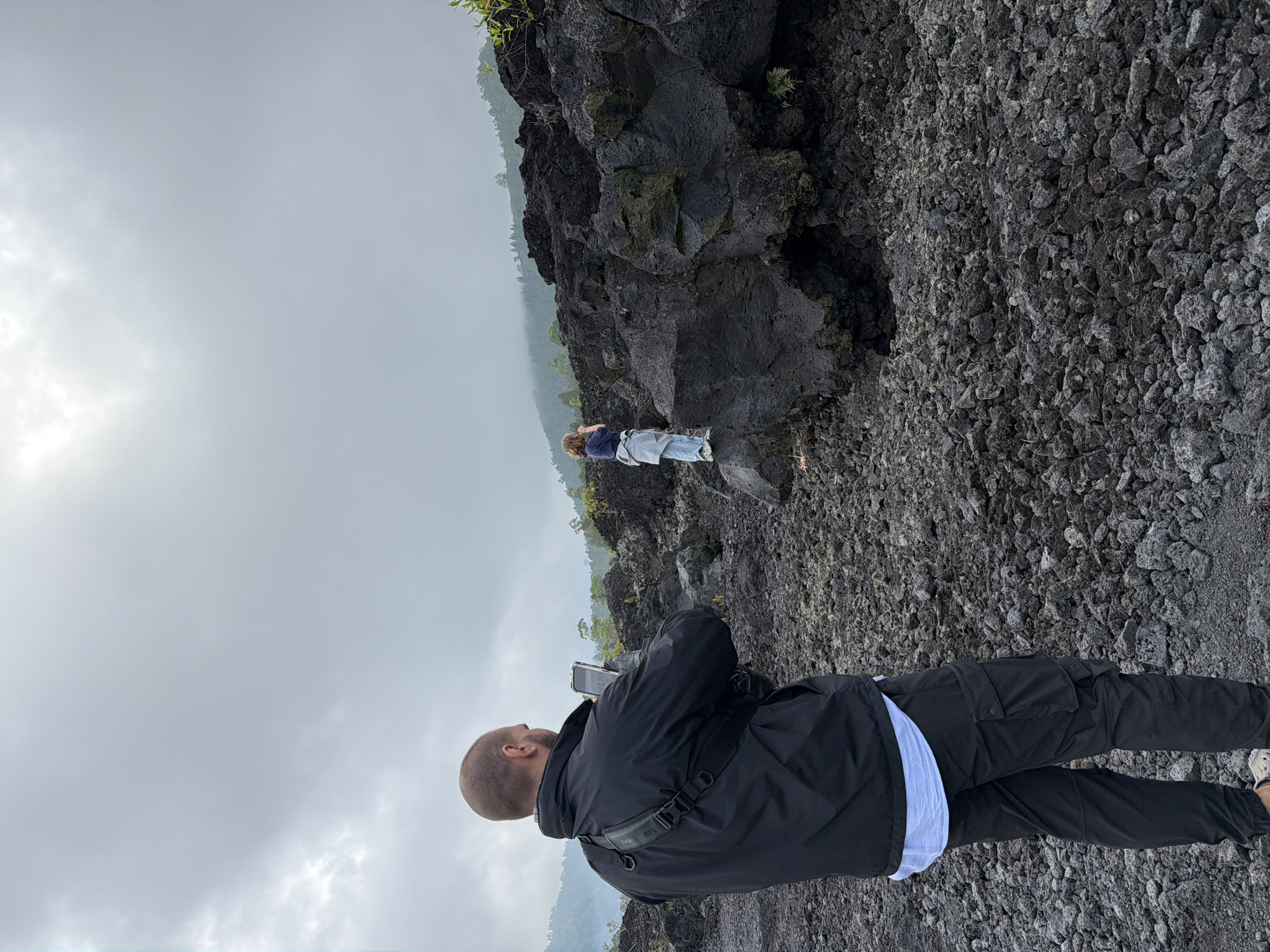 In Kintamani, Indonesia, two individuals are present in a rocky, volcanic landscape. In the foreground, a bald man wearing a black jacket, a light blue shirt, and black cargo pants is holding a smartphone to take a picture. He is positioned with his back to the camera, looking towards a person standing on a dark, porous rock formation in the mid-ground. This second person, seen from behind, is wearing a blue shirt, a grey jacket tied around their waist, and light-colored pants. They are also holding a phone, presumably taking a photo or video of the surrounding environment. The ground is covered in dark, irregular volcanic rocks and stones, with sparse vegetation visible on the rocky slopes and in the background. The sky is overcast with a thick layer of clouds, and a misty haze obscures the distant hills and trees. The overall scene suggests a visit to a volcanic site, possibly an active or recently active one, given the stark, rocky terrain.