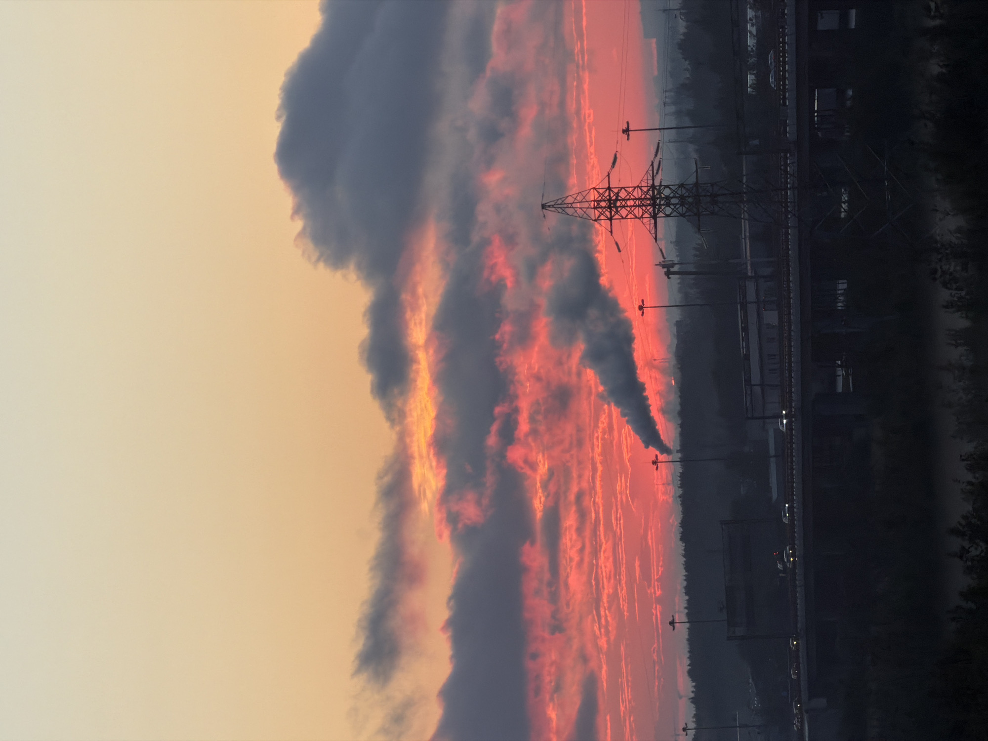 The image captures an industrial landscape at dusk, likely from an elevated vantage point. A vibrant sunset paints the sky with hues of orange, yellow, and deep red, illuminating a dramatic display of clouds. Plumes of smoke rise from industrial chimneys, adding to the atmospheric conditions. A tall, latticed power pylon stands prominently in the foreground, silhouetted against the colorful sky. Below, a multi-level highway is visible, with cars illuminated by their headlights traversing the dark roadway. In the background, a dense line of trees forms a dark horizon, obscuring the details of the city beyond. The overall impression is one of industrial activity juxtaposed with the natural beauty of a sunset.