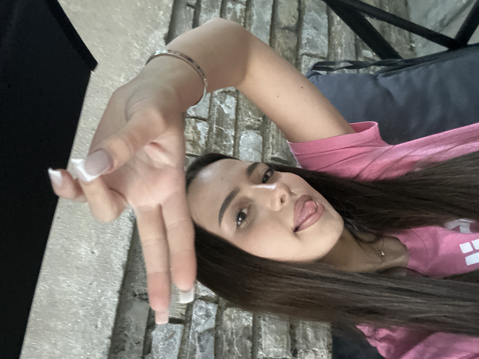 A woman lies at an angle, wearing a pink shirt. Her right arm is raised, and her hand is making a gesture. She has long brown hair, light skin, and is sticking her tongue out slightly. She is wearing a silver bracelet on her right wrist. A portion of a black object is visible in the upper left corner. She is sitting on a gray cushion. The background appears to be a stone wall. The location is Unknown, Unknown.