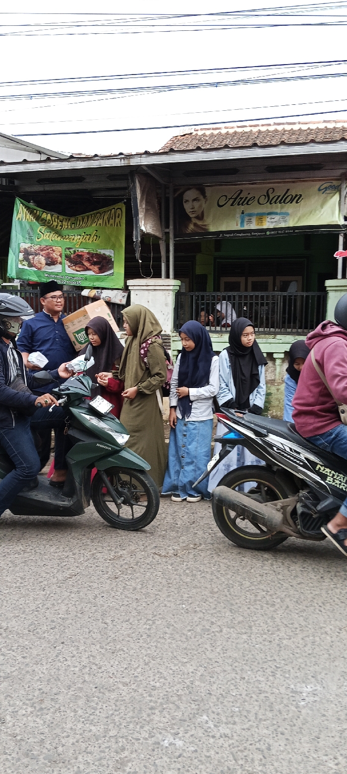 The image shows a street scene in Cangkuang, Indonesia, located at latitude -7.065 and longitude 107.552. The scene depicts a group of people standing and interacting in front of a shop. A man is wearing a black shirt and a black skullcap and is holding a small box. He is standing next to a man with a blue shirt and is holding a white paper bag in his left hand. Behind them is a dark green Honda motorbike and a man is driving it, wearing a blue shirt and black helmet. There is another motorcycle parked next to the Honda motorbike. Behind the Honda motorbike is a woman in a long green gown. Behind her is a woman in a light blue shirt and a long blue skirt. Behind her is a woman in a black hijab and a black shirt, and a woman in a black hijab and a white shirt. The woman in a black hijab and black shirt is looking to her left, and the other woman is looking to the right. The woman in a black hijab and a white shirt is holding a brown bag on her right shoulder. There is another woman in a grey shirt and a black hijab. There is a sign on the wall that says "Arie Salon." The sign is in Indonesian, which is the official language of Indonesia. There is another sign in Indonesian. The street is paved with concrete. There are power lines in the sky above the buildings. There are a few palm trees in the background. There is a large metal gate behind the people in the scene. The day is overcast, with the sky a light grey.