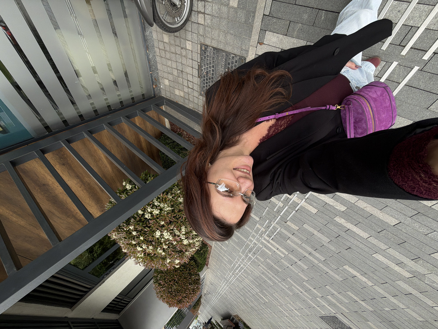 This selfie captures a smiling woman in an urban outdoor setting in Belgrade, Serbia. The image is taken from a slightly high angle, with the woman looking up towards the camera.
The woman has long, dark brown hair that cascades over her shoulder. She wears clear-rimmed glasses and a warm smile. Her outfit consists of a dark, possibly black, blazer or coat worn over a deep burgundy or plum-colored top, which appears to have a lace trim peeking out. She carries a vibrant, suede-like purple cross-body bag with gold hardware. Light-colored pants, likely jeans, and dark red or burgundy shoes are visible on her lower body.
The background reveals a modern urban environment. To the left, part of a building features vertical frosted glass panels and dark gray metal railings. Behind these railings, wooden slats are visible, alongside lush green bushes, some adorned with small white flowers. The ground is paved with rectangular gray and lighter gray stones arranged in an alternating pattern. A dark manhole cover is partially visible on the ground, and in the far top-left corner, a motorcycle wheel is slightly visible.
The lighting suggests it is daytime, possibly overcast or with diffused sunlight, creating soft shadows. The woman's relaxed posture and smile convey a pleasant and casual mood. No legible text is visible in the image.