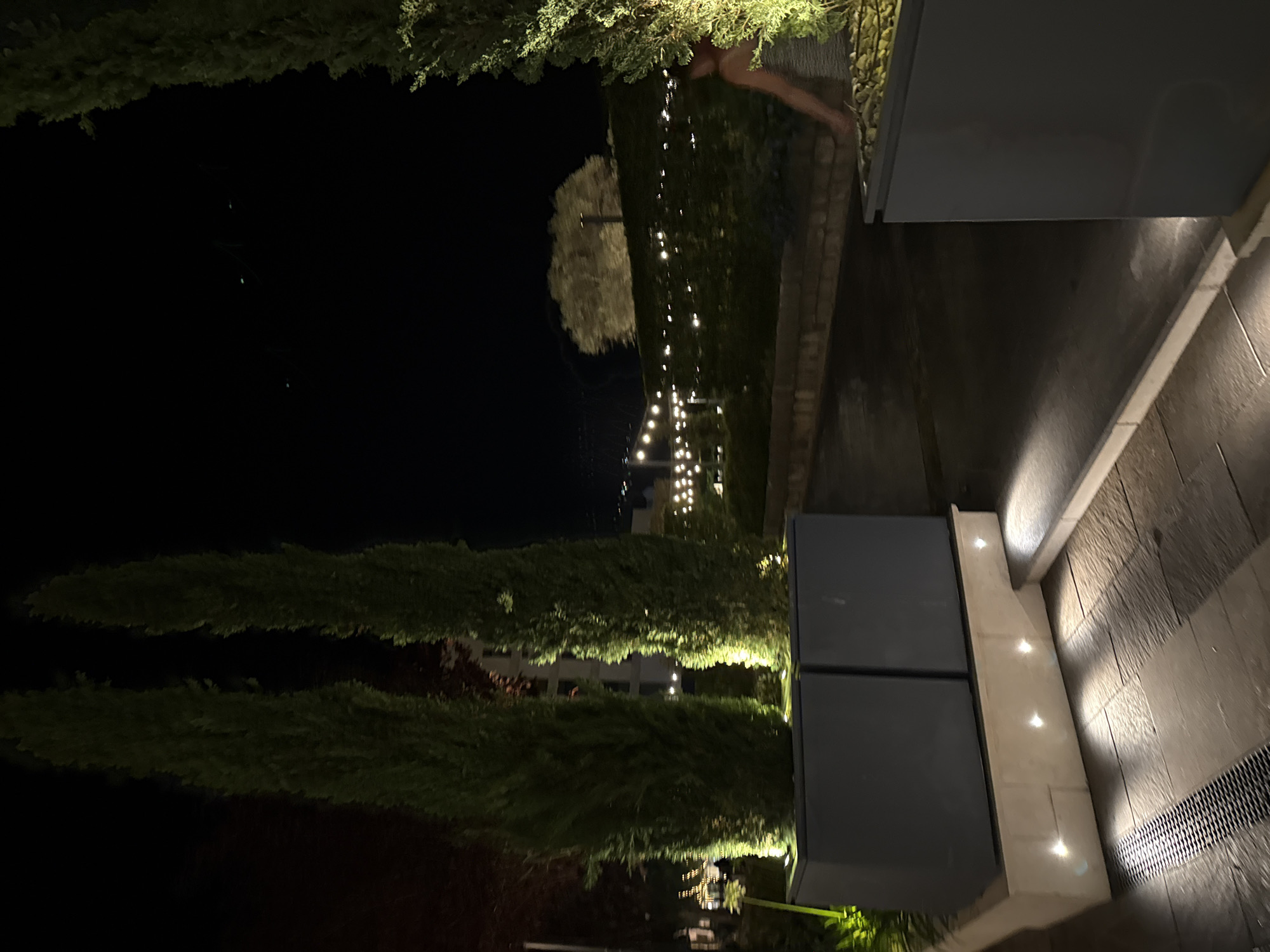 The image depicts an outdoor scene at night, possibly in a resort or restaurant setting, in Tivat, Montenegro. In the foreground, two tall, slender, dark green cypress trees stand prominently within large, rectangular, dark grey planters. These planters are elevated on a light-colored stone base, which is illuminated by several small, warm-toned lights embedded in its front. The ground in front of the planters is paved with light-colored stone tiles, with a dark metal grate visible on the left side.
In the mid-ground, a pathway or paved area extends into the distance. Along the sides of this pathway, there are lush green hedges. A string of small, warm-toned lights hangs above the hedges, creating a festive atmosphere. Further in the background, a large, light-colored tree is visible, illuminated from below. A person, appearing to be a woman with dark hair and wearing a striped garment, is partially visible on the right side of the image, with their back to the camera, near another dark grey planter. The overall scene suggests an elegantly lit outdoor area designed for evening ambiance. The sky above is dark and clear, indicating nighttime.