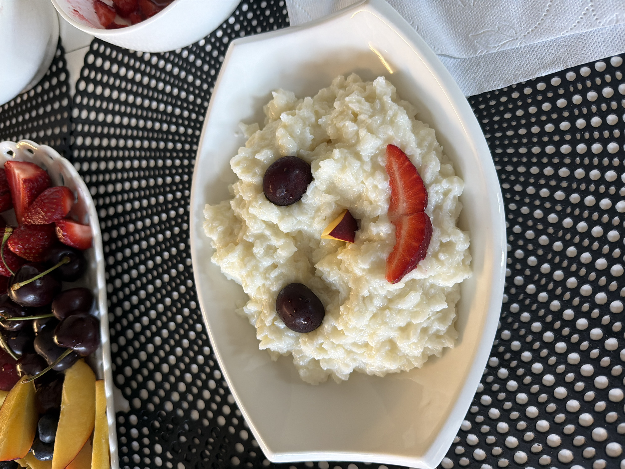 A white, multi-faceted ceramic bowl is centrally positioned, containing a serving of creamy white rice pudding. The pudding is artfully decorated to form a simplified face: two dark red cherries serve as eyes, a small triangular piece of yellow-orange fruit, possibly peach or apricot with a reddish edge, forms the nose, and a curved slice of bright red strawberry represents a smile.
In the upper left background, a white decorative platter is laden with an assortment of fresh fruits, including slices of yellow peaches or nectarines, clusters of dark red cherries with stems, blueberries, and halved strawberries. To the upper right, a partially visible white bowl contains additional sliced strawberries. A crumpled white paper napkin lies to the immediate right of the central bowl.
All items rest on a black placemat featuring a uniform pattern of small white circular dots. The surface beneath the placemat appears light in color. The scene is brightly and evenly lit, indicating an indoor environment. No individuals are visible in the frame.