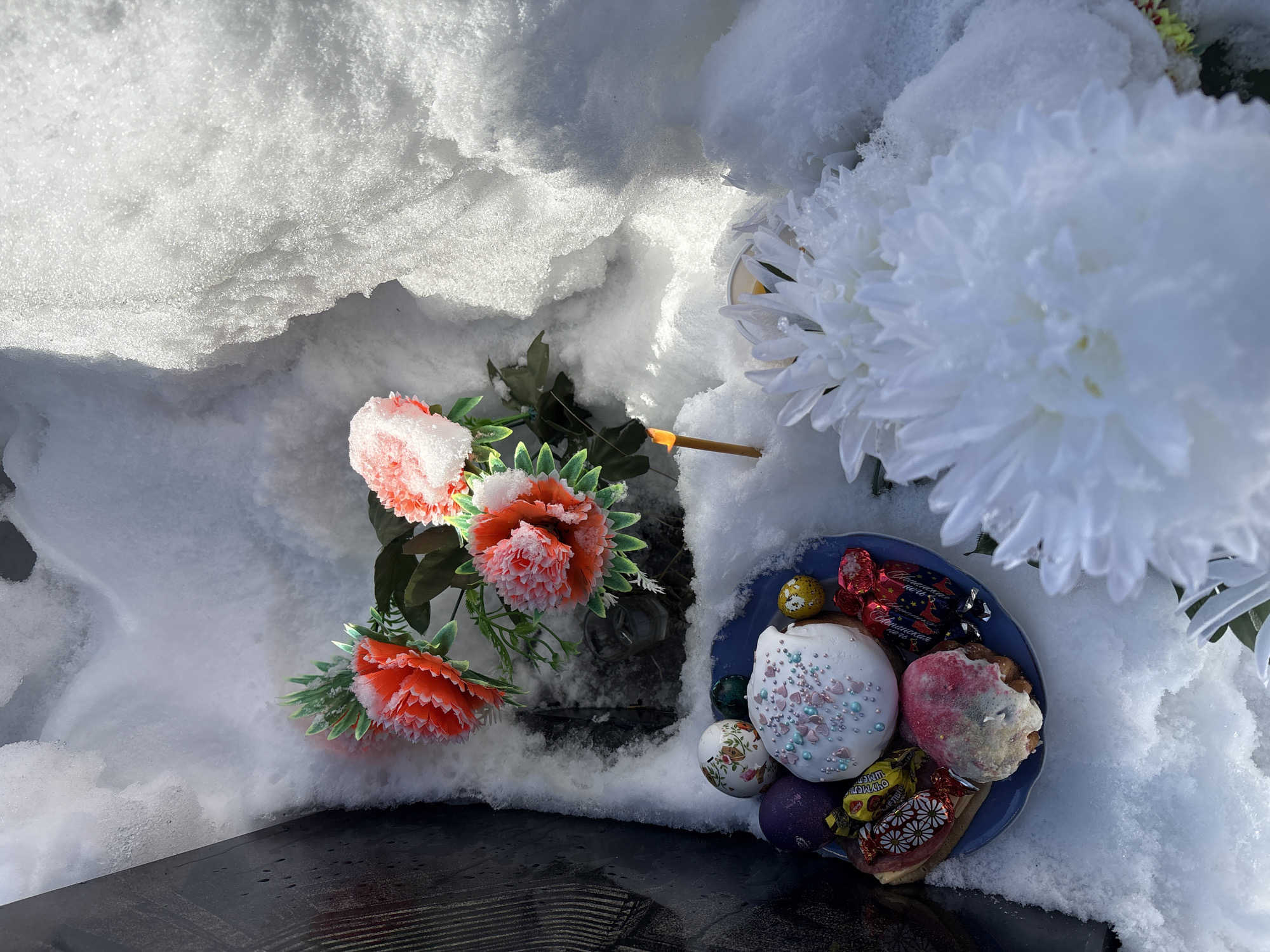 This outdoor image depicts offerings made on a snow-covered surface, strongly suggesting a memorial or grave site. Much of the frame is dominated by bright, granular snow, sparkling under direct sunlight and casting soft shadows.
On the left side, a cluster of artificial flowers, primarily in shades of pink and coral with green leaves, is visible, their petals lightly dusted with snow. To the right, a large, artificial white chrysanthemum-like flower also peeks out from the snow. Near the center, a small yellow candle or candle fragment stands upright in the snow.
In the bottom right corner, a dark blue plate holds several items. These include two small, decorated cakes or pastries; one with white frosting and pink and blue heart sprinkles, and another with red/pink frosting. Alongside these are several individually wrapped candies in colorful foils, and at least two decorated eggs, characteristic of Easter eggs—one patterned and one solid purple. Visible text on a red candy wrapper reads "Вдохновение Шоколадные конфеты" (Inspiration Chocolate candies). Other wrappers show partial text like "От Метро" (From Metro) and "ЯШКИНО" (YASHKINO).
A dark, reflective surface, possibly a memorial slab or a tombstone, is visible at the very bottom of the frame, partially covered by snow. The presence of snow, combined with the traditional offerings of Easter cakes, decorated eggs, candies, and flowers, points to a commemoration of the deceased, likely around the time of Easter or Radonitsa (a day of remembrance in Eastern Orthodox Christianity), in Nevon, Russia. The scene conveys a sense of solemn remembrance in a cold but sunny environment.