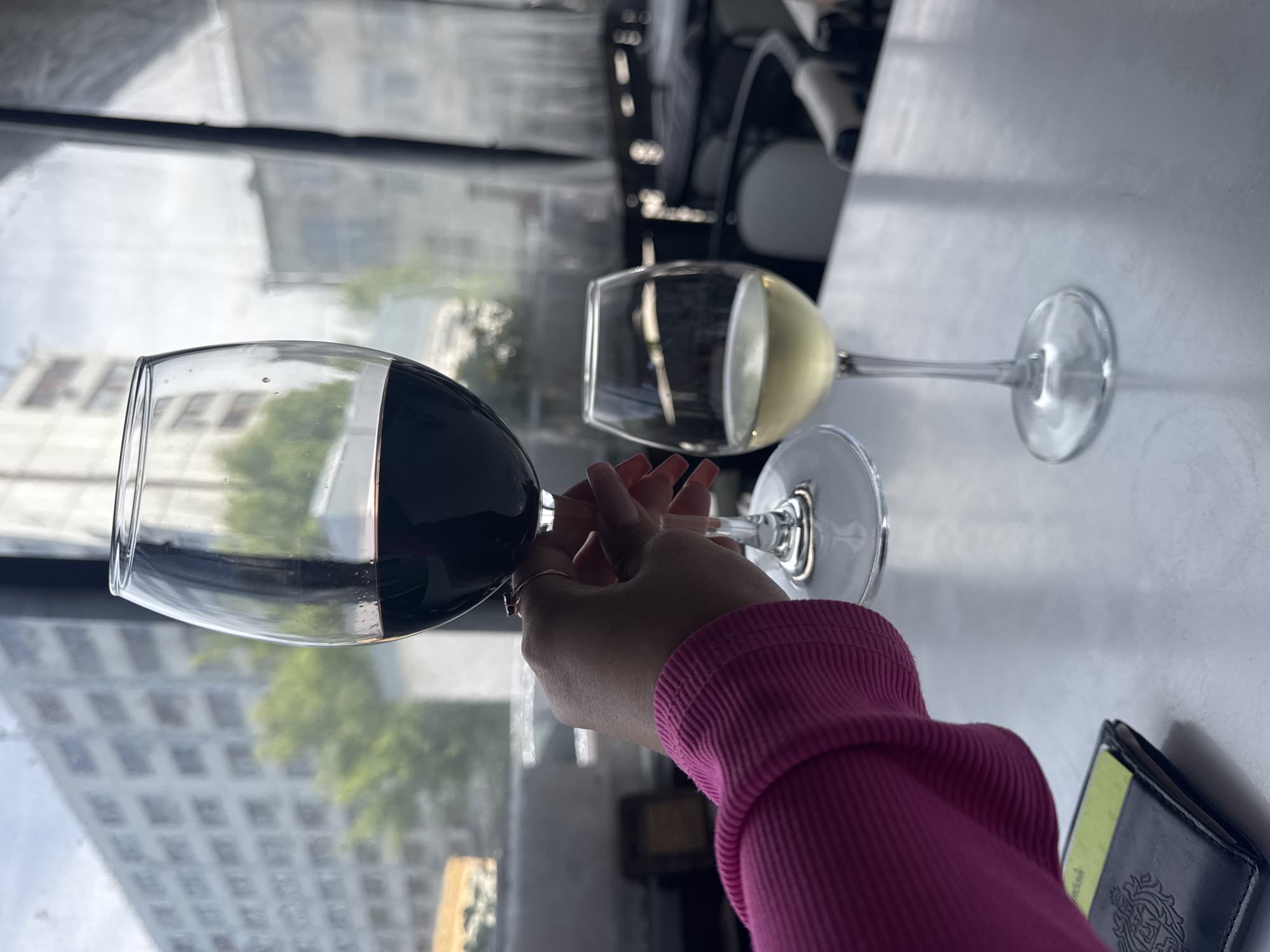 The photograph captures an intimate indoor scene, likely within a cafe or restaurant, overlooking an urban environment through a large window. In the foreground, a person's hand, adorned with long, light-colored nails, holds a glass filled with red wine. The individual is wearing a vibrant fuchsia or magenta ribbed long-sleeved top.
On the light-colored table next to the hand, a second wine glass, containing white or pale yellow wine, is visible. Through the window, a multi-story building with numerous windows is visible, partially obscured by green foliage from trees. The diffused lighting suggests it is daytime, possibly with an overcast sky. To the right of the person's arm, a dark wallet or passport holder rests on the table, featuring a faint, unreadable emblem or crest. The scene conveys a sense of relaxation or social enjoyment in an urban setting, consistent with the provided location context of Chaikovsky, Russia.