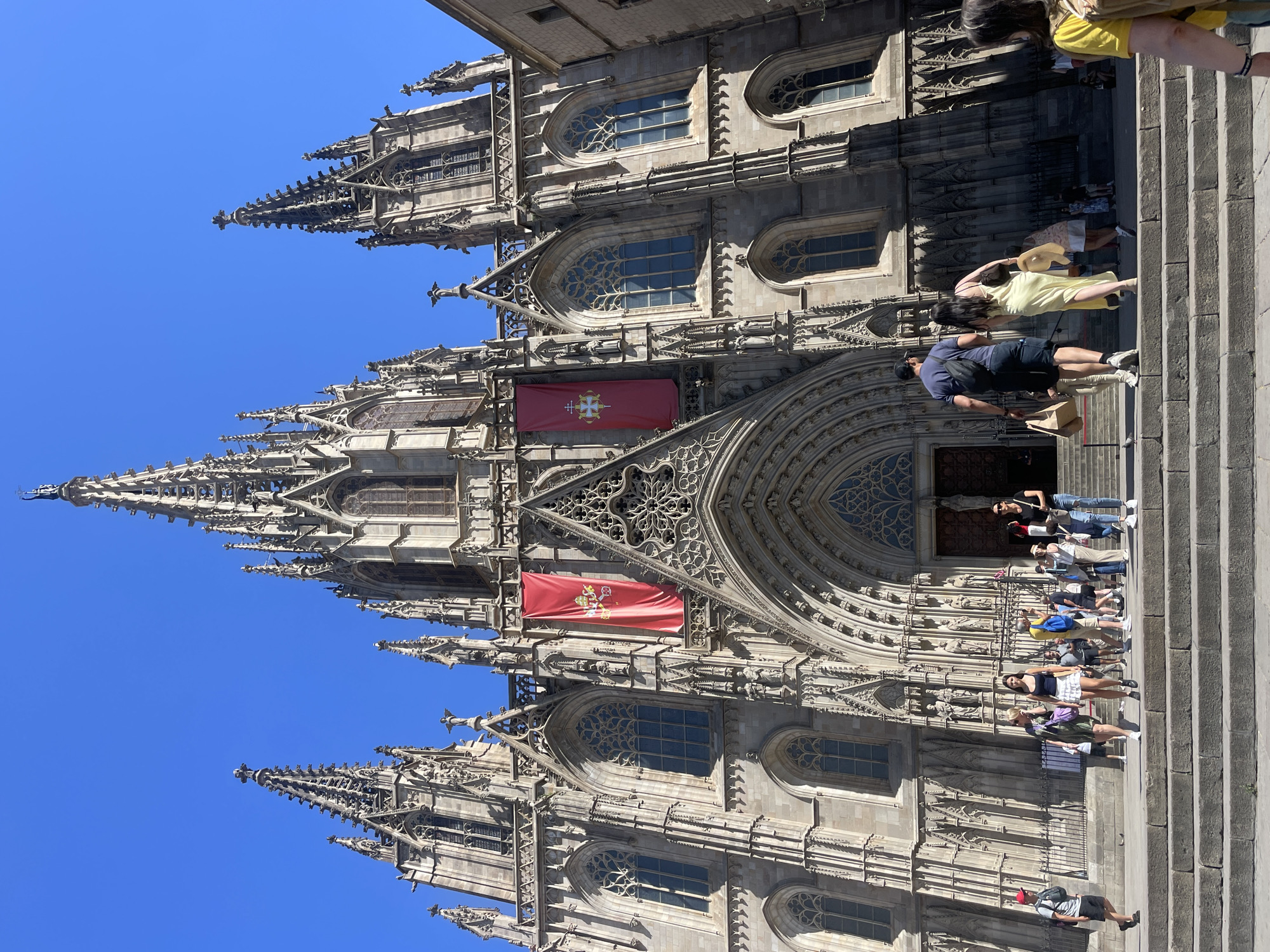 This is a low-angle, full-shot photograph of the Barcelona Cathedral, also known as the Cathedral of the Holy Cross and Saint Eulalia, located in the Gothic Quarter of Barcelona, Spain. The cathedral is a magnificent Gothic structure with intricate stonework, towering spires, and large stained-glass windows. Two large red banners with a golden crest are hanging on the facade. The sky is clear and blue. In the foreground, a set of stone stairs leads up to the cathedral entrance. Several tourists are visible on the stairs and in front of the cathedral, some walking up, others looking around. They are dressed in casual summer clothing. The overall scene depicts a sunny day with tourists visiting a prominent historical landmark.