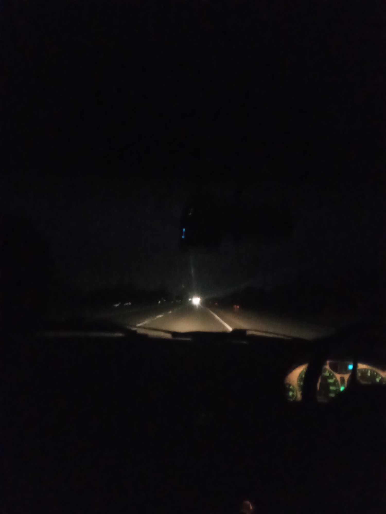 The image captures a nighttime view from inside a vehicle traveling on a highway. The dashboard lights, which appear to be green, are illuminated. The road ahead is visible with lane markings, and other vehicles' headlights and taillights can be seen in the distance. The overall scene is dark, suggesting a rural or suburban area outside the city of Almaty, Kazakhstan. There are no people clearly visible within the frame.
