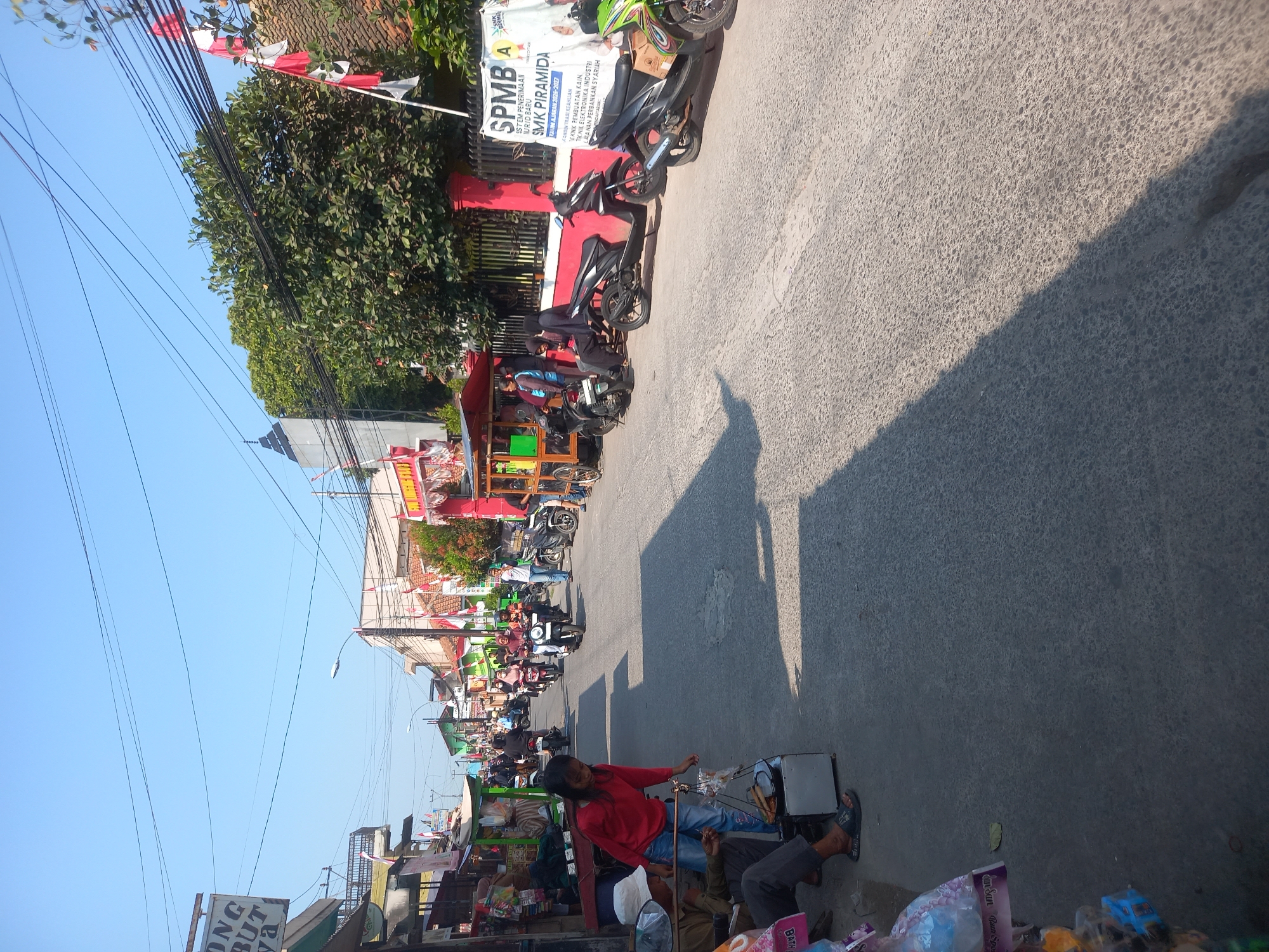 The image depicts a street scene in Linggar, Indonesia, on a clear, sunny day. The street is lined with buildings and market stalls, with several motorcycles parked along the side. A few people are visible, some walking, others sitting or standing near the stalls. In the foreground, a young girl in a red shirt is interacting with an elderly person sitting on the ground next to a food cart. Red and white Indonesian flags are displayed on some of the buildings, suggesting a national celebration or event. The overall atmosphere appears to be lively and bustling, characteristic of a local market area.