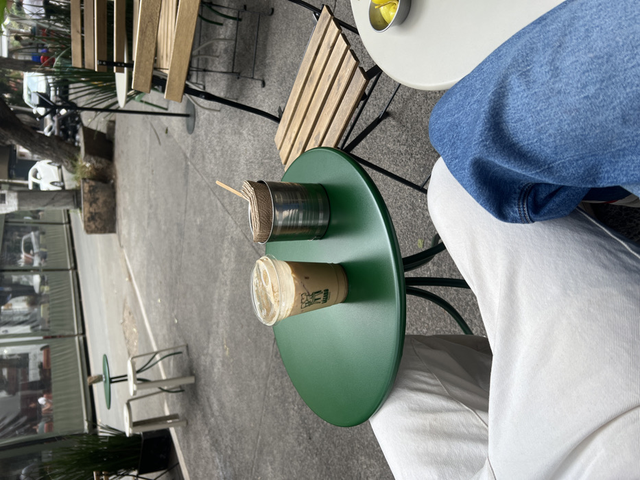 The scene depicts an outdoor cafe setting on a sidewalk in Mexico City, Mexico. In the immediate foreground, the legs of two individuals are visible: one wearing white pants on the left, and another wearing blue denim jeans on the right. A round, dark green metal table occupies the central foreground. On this table, a clear plastic cup containing iced coffee with foam and a green "LAZARO" logo is present, alongside a cylindrical metal can holding several folded brown paper napkins and a wooden stir stick. To the right, a portion of a light-colored round table is visible, featuring a small metal bowl filled with yellow citrus wedges. A wooden slat chair with a black metal frame is partially visible behind this table. The background shows a grey concrete sidewalk bordering a street with blurred vehicular traffic, including a white car and a motorcycle. Trees line the street, and a building with an outdoor dining area, enclosed by a clear plastic barrier, is discernible across the street. Additional outdoor seating, including small tables and stools, is visible further down the sidewalk.