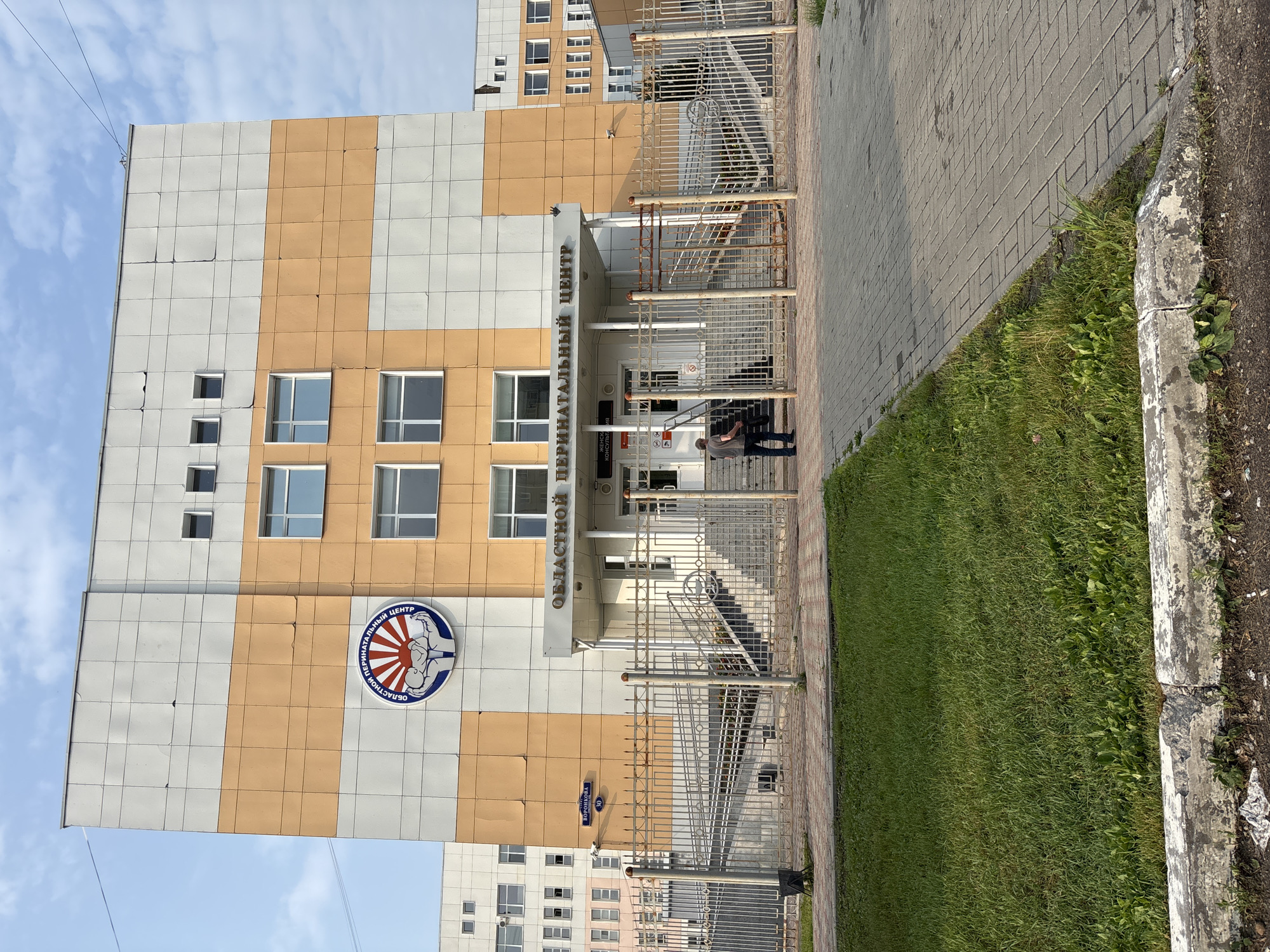 The image shows the front of the Blagoveshchensk Regional Perinatal Center, located at 30 Voronkova Street in Blagoveshchensk, Russia. The building is multi-story with a facade of alternating orange and light grey panels. A circular emblem featuring stylized hands cradling a baby, with sun rays emanating from behind, is mounted on the upper part of the building. The words "ОБЛАСТНОЙ ПЕРИНАТАЛЬНЫЙ ЦЕНТР" (Regional Perinatal Center) are inscribed above the entrance. The entrance is accessed via a ramp and stairs, all protected by a metal fence. A single man is standing in front of the fence, looking towards the building. A paved walkway and a grassy area are in the foreground. The sky is blue with some clouds.
