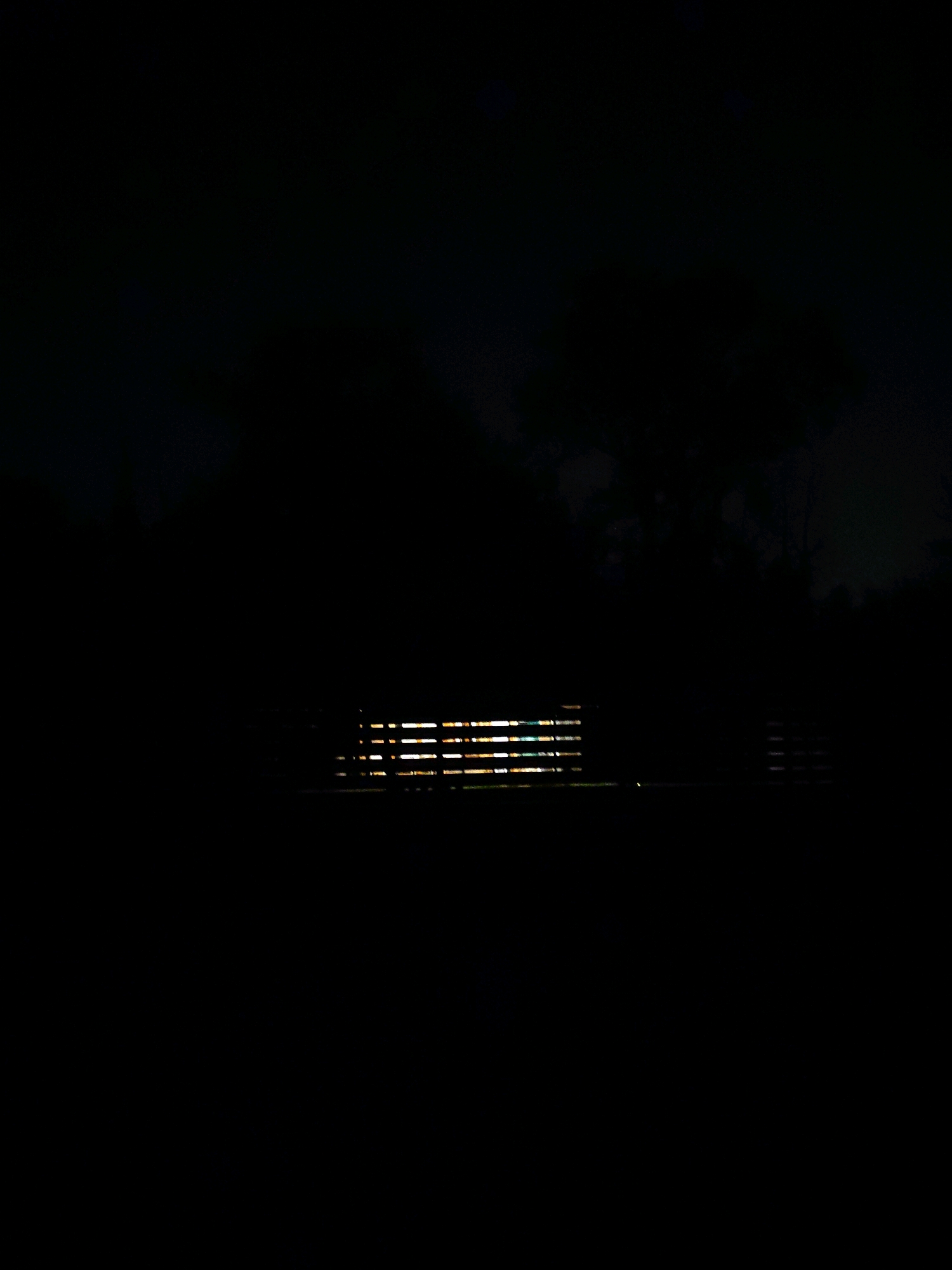 The image shows a dark, outdoor scene at night, with a very low level of ambient light. In the middle of the frame, a long, horizontal structure is visible, illuminated from behind by a series of bright, horizontal lights. This structure appears to be made of parallel slats or bars, suggesting it could be a fence, a bench, or part of a railing. The background is almost entirely black, with faint suggestions of foliage or trees in the upper right corner. The overall impression is one of a dimly lit, possibly secluded, outdoor area.