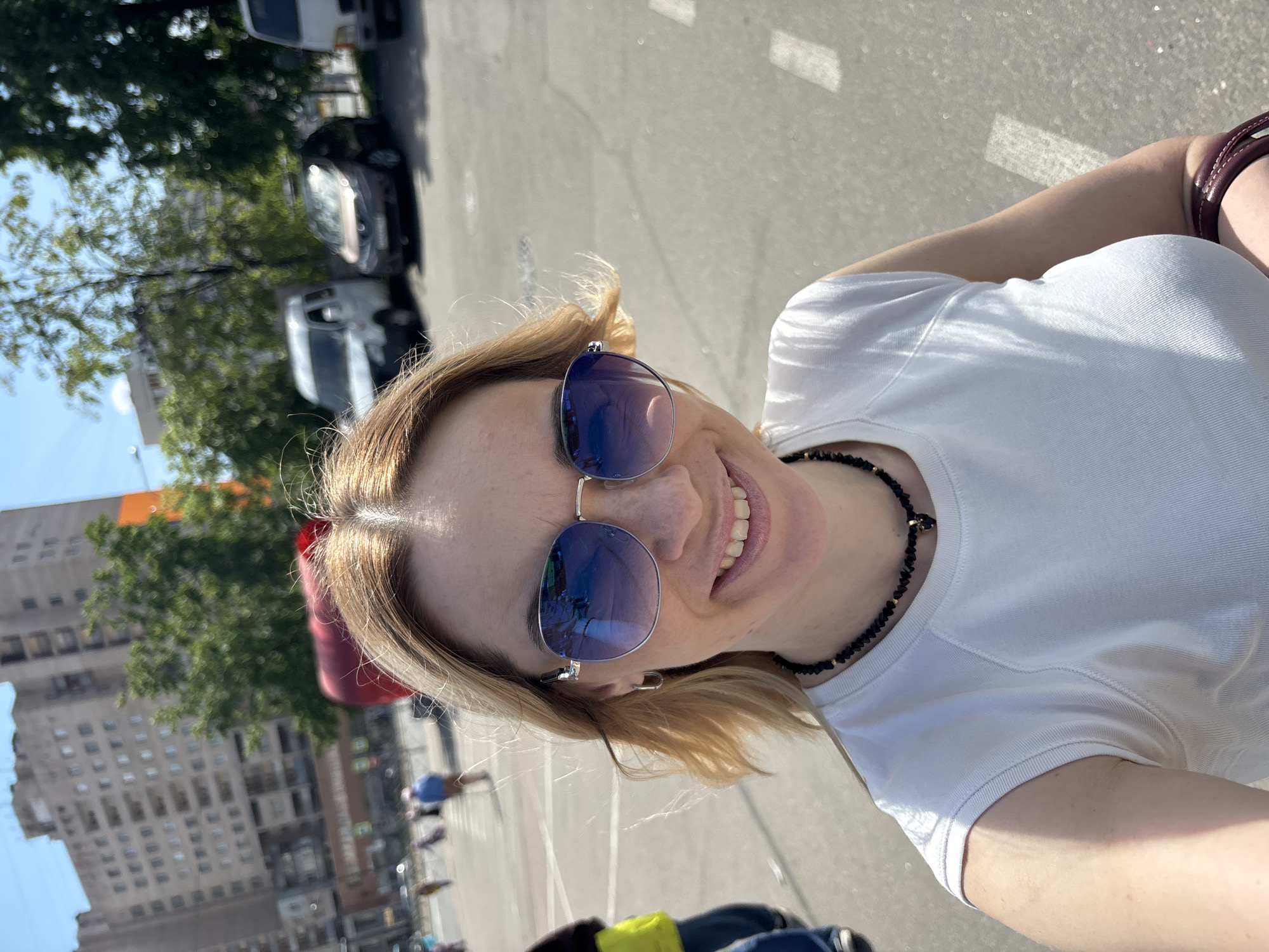 A fair-skinned woman with shoulder-length blonde hair is depicted in a brightly lit outdoor selfie. She is smiling broadly, showing her teeth, and wears large, blue-tinted reflective aviator sunglasses. She is dressed in a white ribbed t-shirt and a black beaded necklace with small gold accents. A dark red or brown handbag strap is visible over her right shoulder.
The background shows an urban environment on a clear, sunny day. The ground appears to be a paved road or parking lot with white painted lines. Several vehicles, including cars and a white van, are visible in the distance. Tall, light-colored buildings and green trees line the street, suggesting a city setting. The bright sunlight creates clear shadows, indicating midday conditions. The overall mood is cheerful and casual. No discernible text is visible in the image.