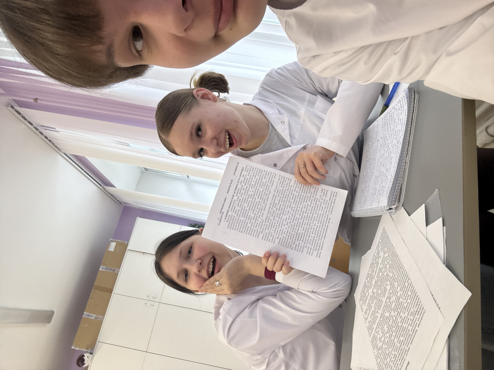 Three young individuals, presumably students, are posing for a selfie inside what appears to be a classroom or laboratory, given their white coats and the presence of papers and notebooks. The classroom setting is indicated by the cupboards and other furnishings visible in the background. One person at the top, cropped. One person with hair tied back in a ponytail is to the right, and a third is slightly below, holding up a paper with text in Russian, suggesting they may be engaged in academic work or research. The location is inferred to be in the city of Chaikovsky, Russia, based on the Russian text.