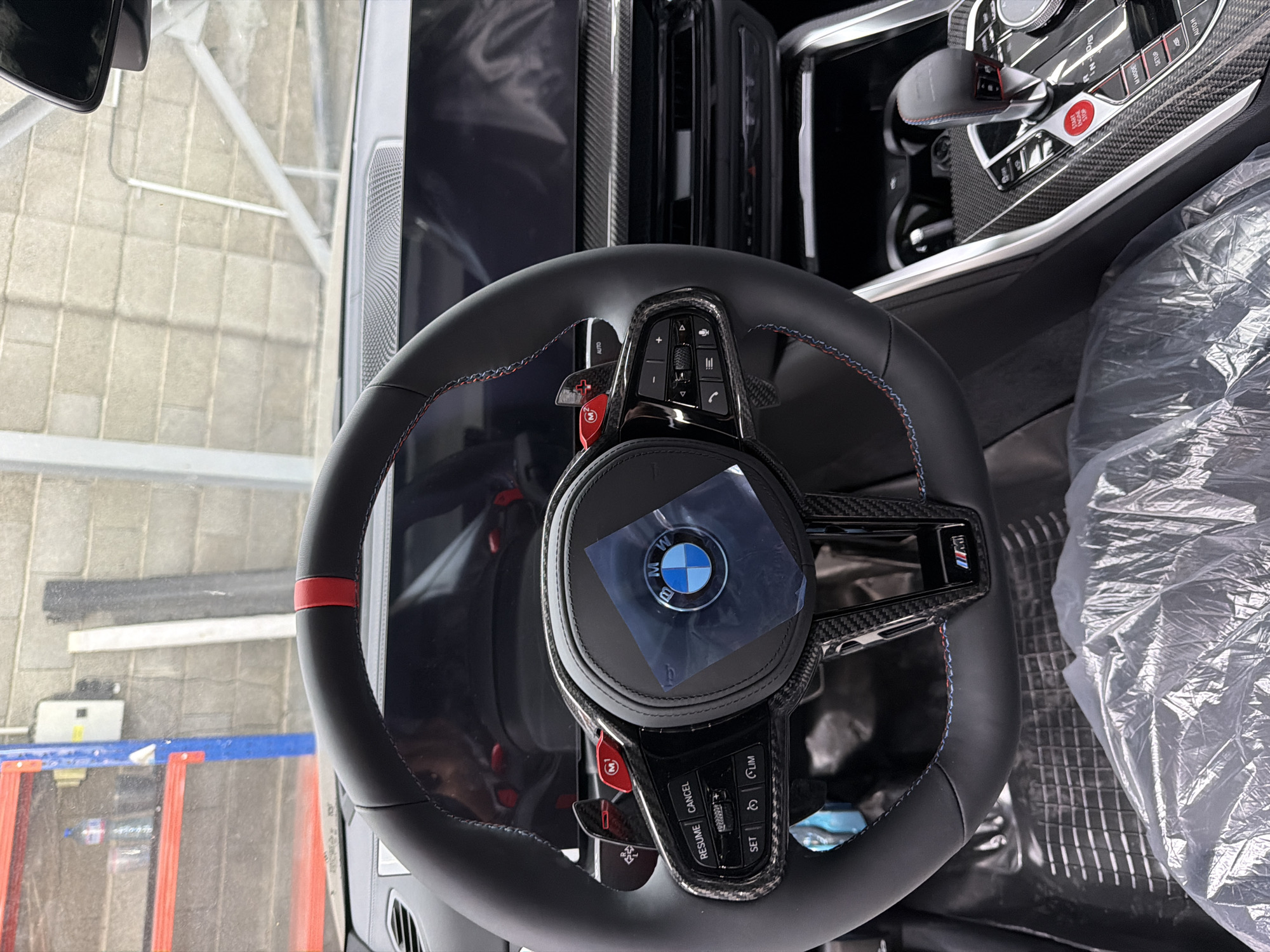 The image shows the interior of a new BMW car, likely a performance model given the M badges and carbon fiber accents on the steering wheel and center console. The steering wheel has red stitching and a red stripe at the top, indicating an M Sport or M model. The center console features the M Mode and Setup buttons, along with a gear selector and the signature red Start/Stop button. The car's interior appears to be covered in protective plastic, suggesting it is either brand new or awaiting delivery. The background shows a garage or workshop environment with shelves, a brick wall, and some equipment. There are no people visible in the image. The specific location in Burgas, Bulgaria, is not identifiable from the visual information provided in the image.
