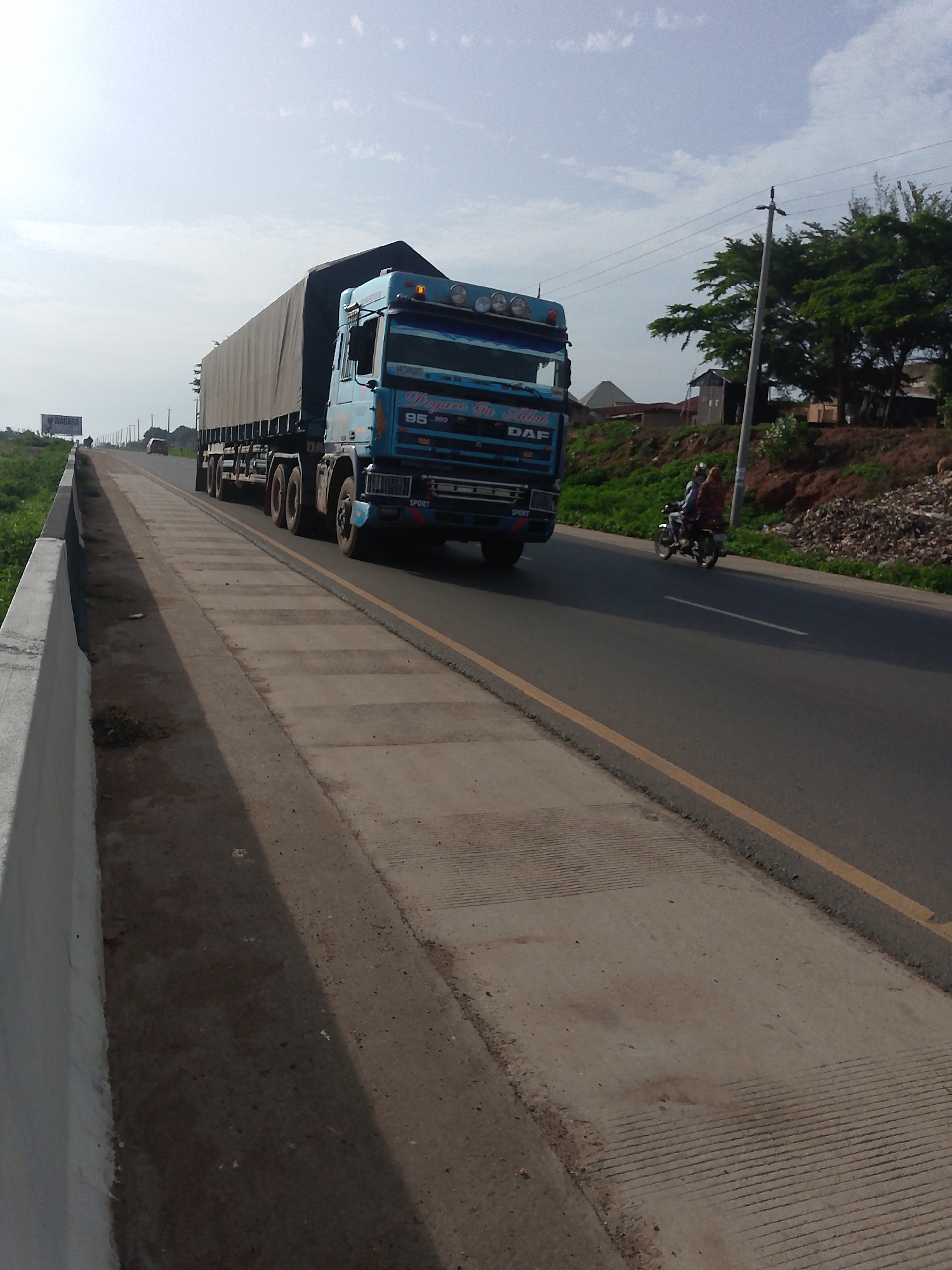 A bright blue DAF semi-trailer truck with a grey tarp-covered trailer is traveling on an asphalt road. The truck has "95" and "360" on its side, along with the text "Sha Allah" and "DAF". The truck has a light brown license plate with white text that appears to be a Nigerian plate. In the right lane, a motorcycle with two occupants is traveling in the same direction as the truck. The driver of the motorcycle is wearing a blue shirt, and the passenger is wearing a patterned brown and red headscarf and shawl. They are traveling on a two-lane road with a yellow line dividing the lanes. The road is bordered on the left by a concrete barrier and a grassy embankment, and on the right by a similar grassy embankment with some trees and buildings visible in the distance. A power pole with wires runs along the right side of the road. In the far distance, a billboard with "WADA" is visible on the left side of the road. The sky is overcast with scattered clouds.
