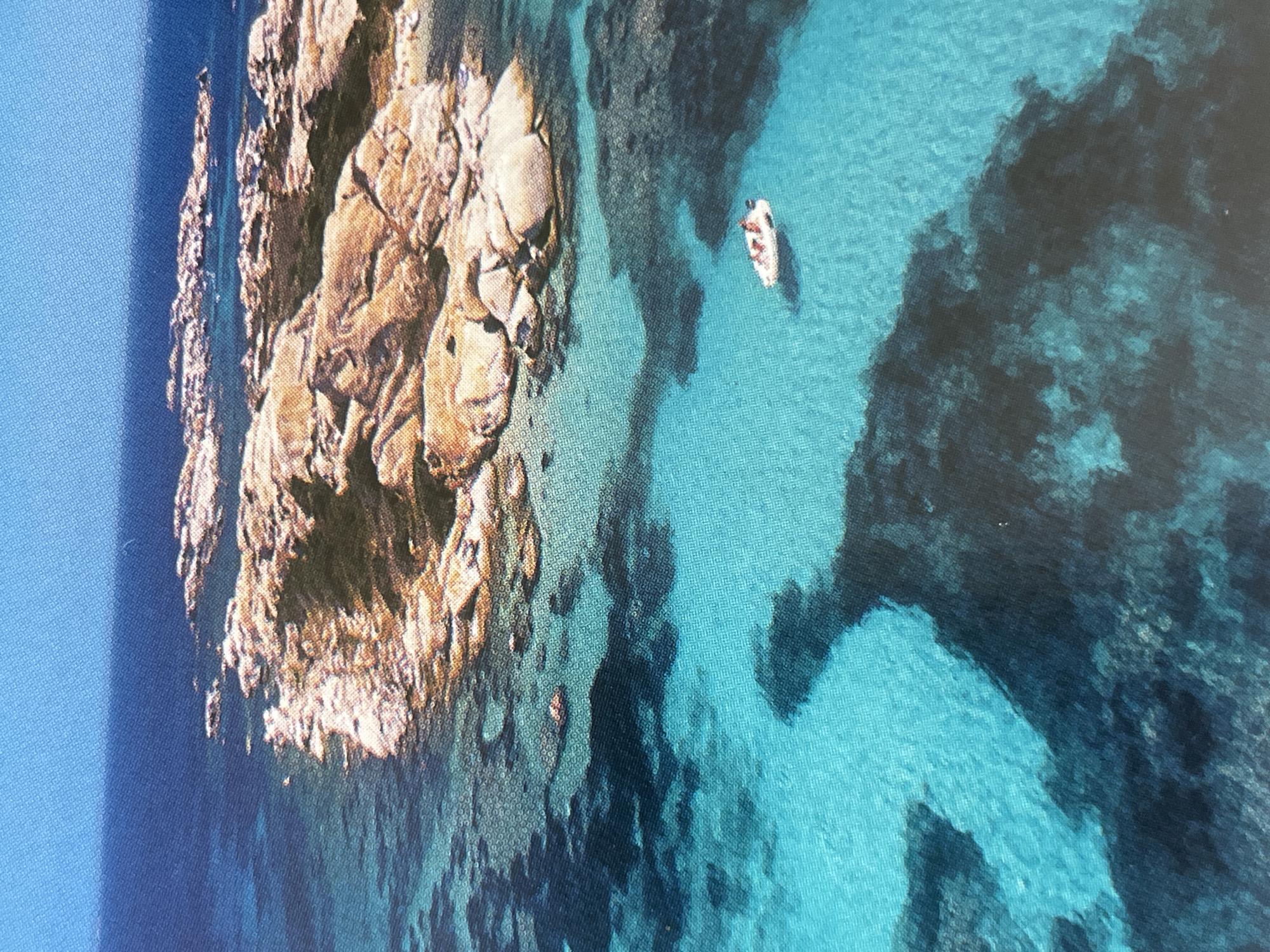 This is an aerial photograph of a rocky island in a clear, turquoise sea. The island is covered with large, smooth, light-brown rocks. The water around the island is shallow and transparent, revealing dark patches of seaweed or coral beneath the surface. A small, white boat with a few people on board is visible in the water to the right of the island. The sky above is a clear, deep blue. The image does not depict any part of the city of Sofia, Bulgaria.
