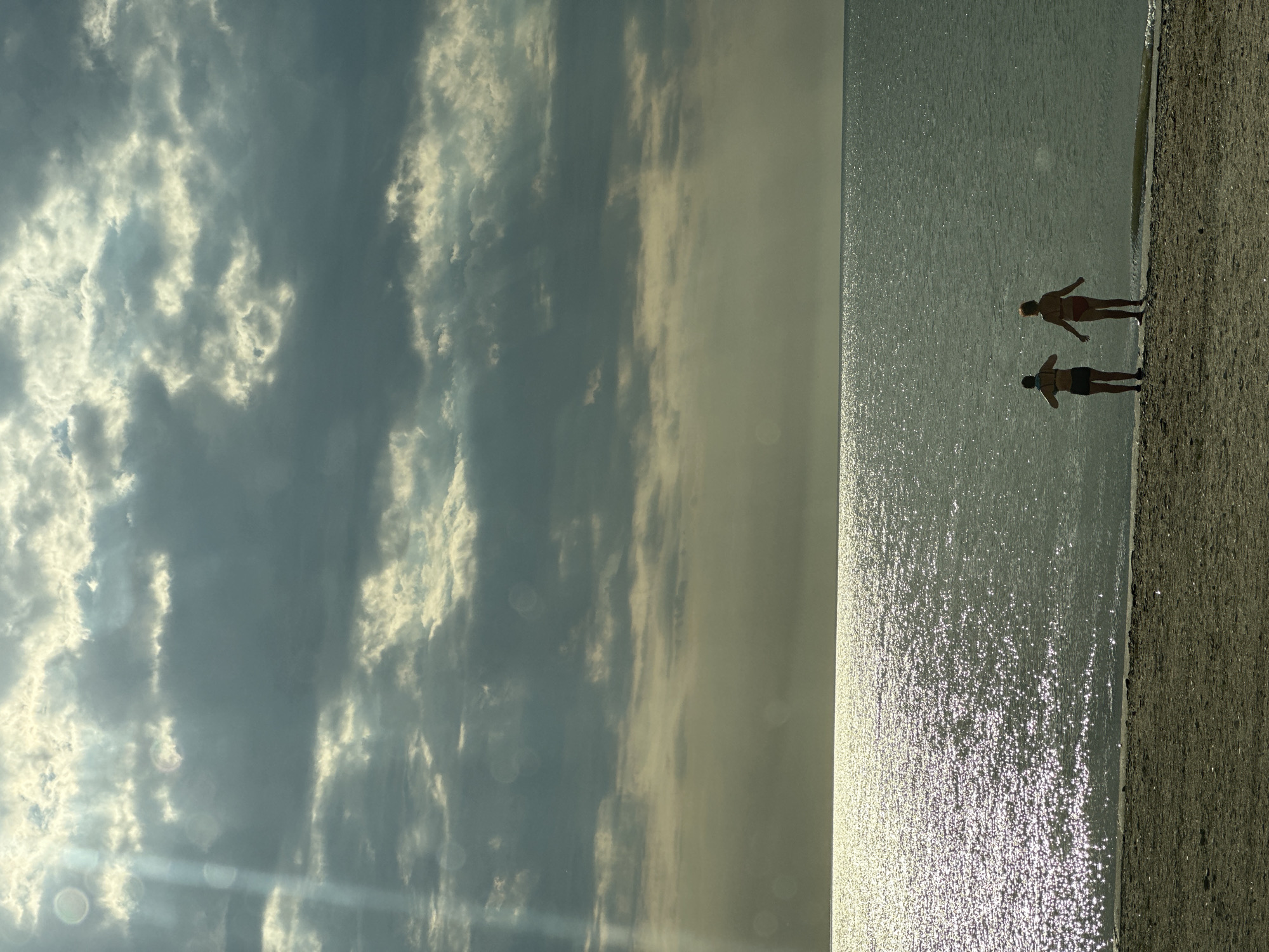 This media file depicts a serene beach scene at what appears to be either sunrise or sunset, given the low angle of the sun and the golden light reflecting off the water. The image is oriented vertically.
In the foreground, on the right side of the frame, is a sandy beach. Adjacent to the beach is a vast expanse of calm water, shimmering with sunlight. Two individuals are visible standing at the water's edge, facing away from the camera and towards the sea. One person, wearing a red bikini, is slightly ahead of the other, who is wearing dark shorts and a dark top. Their posture suggests a moment of quiet contemplation or enjoyment of the view.
The left side of the image is dominated by a dramatic sky filled with clouds. Some clouds are bright and illuminated by the sun, appearing white and fluffy, while others are darker and more diffused, suggesting movement or varying atmospheric conditions. The interplay of light and shadow in the sky creates a sense of depth and atmosphere.
The overall mood of the image is peaceful and tranquil. The setting is clearly a beach or coastline, and the time of day is likely early morning or late evening. The location is identified as Primorsko-Akhtarsk, Russia. There is no visible text in the media file.