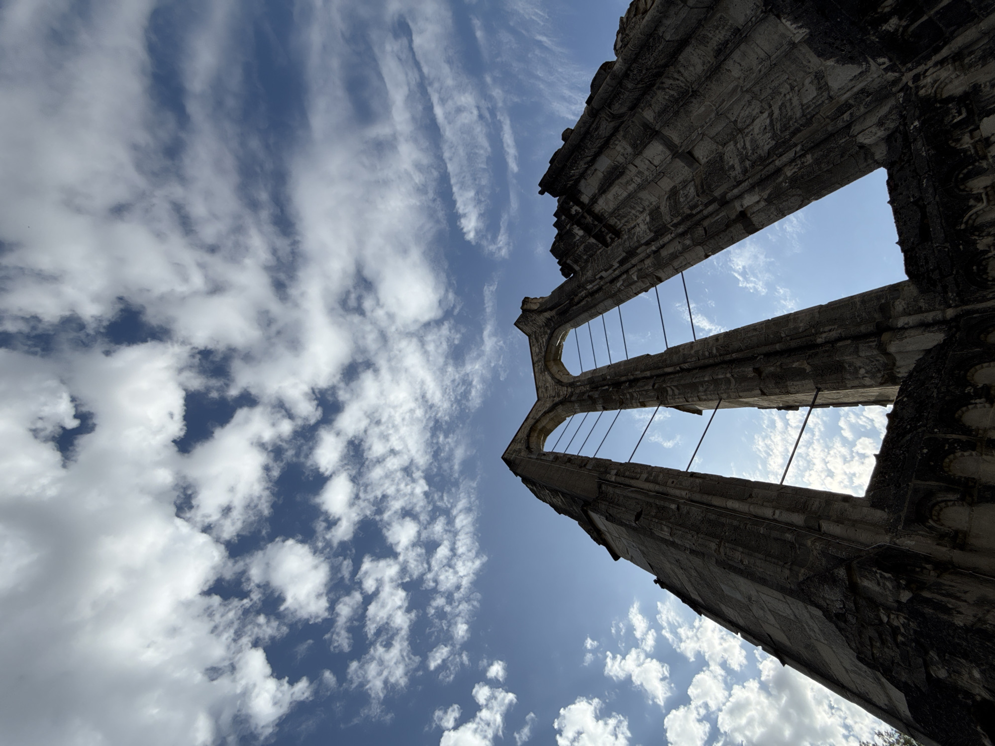 The image captures a low-angle view of the ruins of the Walkenried Abbey in Walkenried, Germany, under a bright, partly cloudy blue sky. The dominant structures are the weathered stone walls of the abbey, with large arched openings that frame the sky. The stone is dark and appears aged, with visible texture and detail in the stonework, including remnants of arched decorations along the lower sections of the walls. Thin, dark metal rods or wires stretch across some of the openings, likely for structural support or as part of the ruins' preservation. The sky is a vibrant blue, with scattered white, fluffy clouds drifting across it. The lighting suggests it is daytime, with the sun casting shadows and highlighting the textures of the stone. The perspective emphasizes the scale and height of the remaining abbey walls against the vastness of the sky.