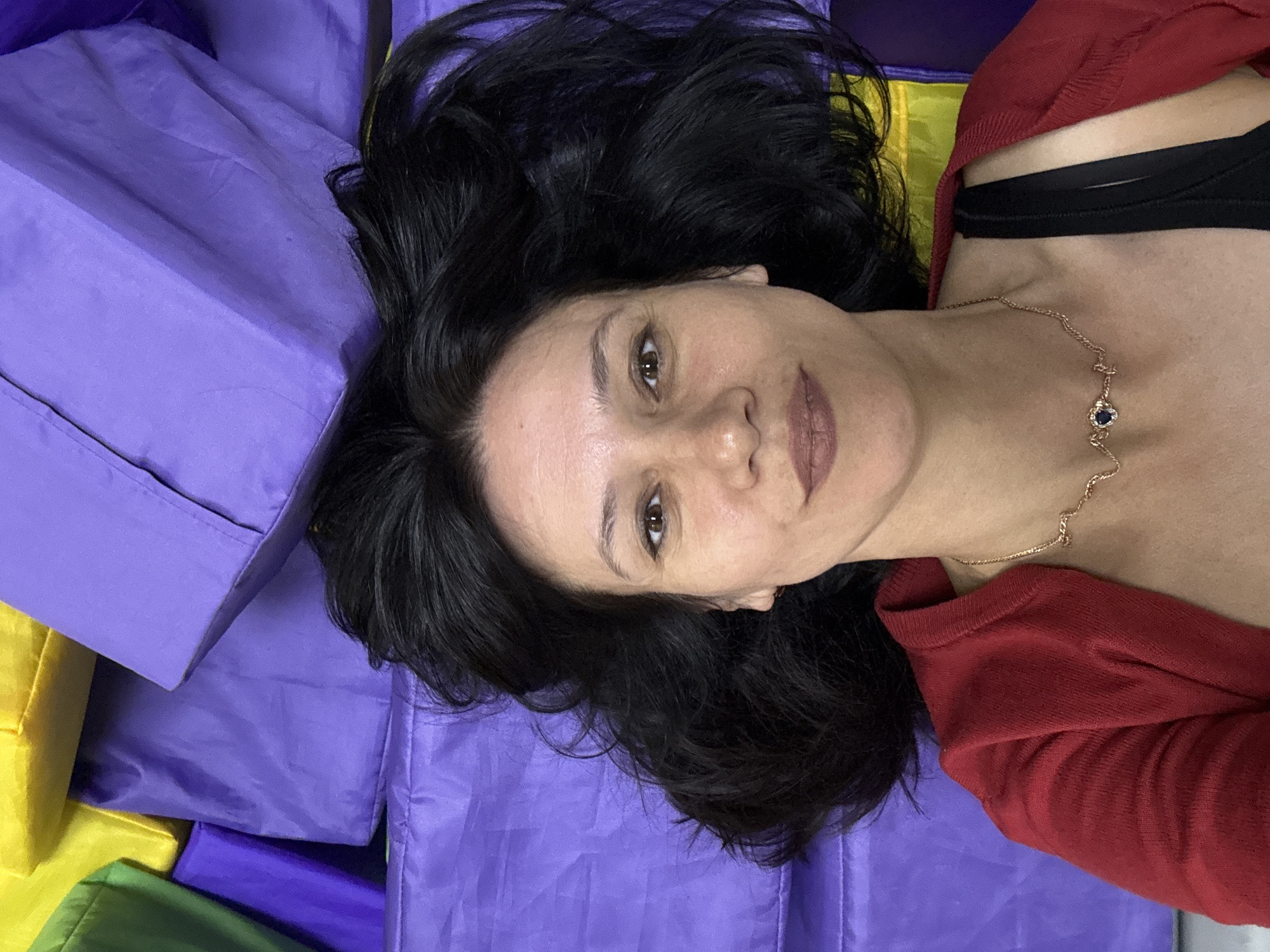 A woman is lying on a surface composed of large, rectangular blocks of varying colors, predominantly purple and yellow. She has long, dark hair spread out around her head. She is wearing a red cardigan over a black bra, and a delicate gold necklace with a dark blue pendant rests on her collarbone. The woman is looking upwards and slightly to the right, with a gentle smile on her face, conveying a sense of relaxation and ease. The lighting is soft, suggesting an indoor environment, possibly a studio or play area, given the presence of the colorful blocks. The overall scene is informal and peaceful. There is no visible text in the image. The context suggests this photo might have been taken in Chaikovsky, Russia, adding a specific geographical element to the setting.