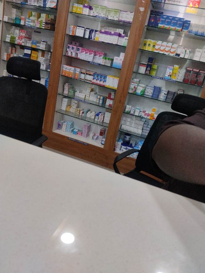A pharmacy interior with a counter in the foreground. Shelves behind the counter are stocked with a variety of medications. Two black chairs are positioned near the counter, one appearing to be occupied. The shelves are organized with different drugs, vitamins, and supplements. The scene is well-lit, reflecting off the glossy counter surface.