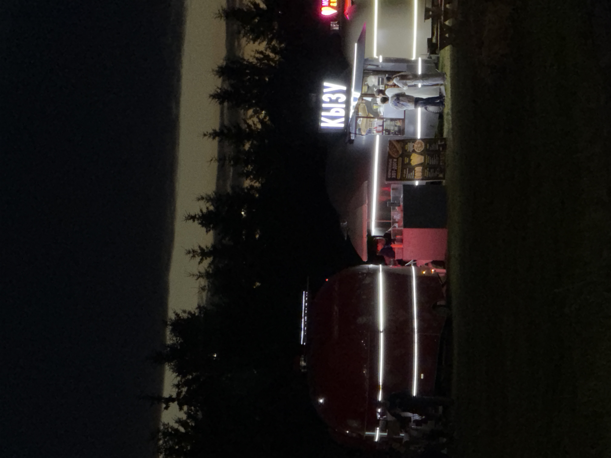 This is a nighttime outdoor scene at a food truck park. The scene is dark with artificial lighting illuminating the food trucks and surrounding areas. In the foreground, a large, red, rounded food trailer with horizontal white LED lights is parked on the left. To the right of the trailer, a smaller food stand with a white canopy and bright LED strip lighting is set up. A sign above this stand displays the Cyrillic word "КЫЗУ" in white illuminated letters. Several people are visible near the food stand, some appearing to be customers or staff. A menu board is mounted on the side of the stand. In the background, a dark silhouette of pine trees lines the horizon under a faintly lit sky. Picnic tables with benches are visible to the right of the food stand, suggesting a seating area. The ground appears to be grassy.