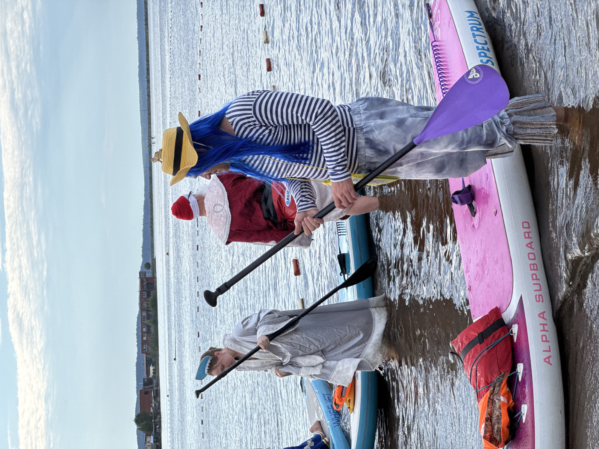 The image shows a group of people on paddleboards in the water. The scene is outdoors, with a cloudy sky and buildings visible in the background. The people are dressed in costumes, including a person with blue hair and a straw hat, another in a Santa Claus outfit, and a third in a gray costume with a white fur trim. They are holding paddles and appear to be participating in an event or activity on the water. One paddleboard has the text "SPECTRUM" and "ALPHA SUPBOARD" visible. A red and orange life jacket is strapped to the pink paddleboard. The water is choppy, suggesting a breezy day. The overall mood seems to be playful and festive, given the costumes. The time of day appears to be daytime, likely afternoon or evening, judging by the light. The location context of Chaikovsky, Russia, provides geographical information about the setting.